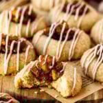 A close-up of apple pie cookies with a focus on one cookie that has a bite taken out, revealing a soft, tender interior. Each cookie is topped with a rich caramelized apple filling and drizzled with a cinnamon-spiced glaze, enhancing its warm, sweet flavor. The cookies are displayed on a wooden board, surrounded by others, capturing their inviting, homemade texture. The vibrant apple filling and glaze drizzle add an autumnal charm, making these cookies look as comforting as they are delicious.
