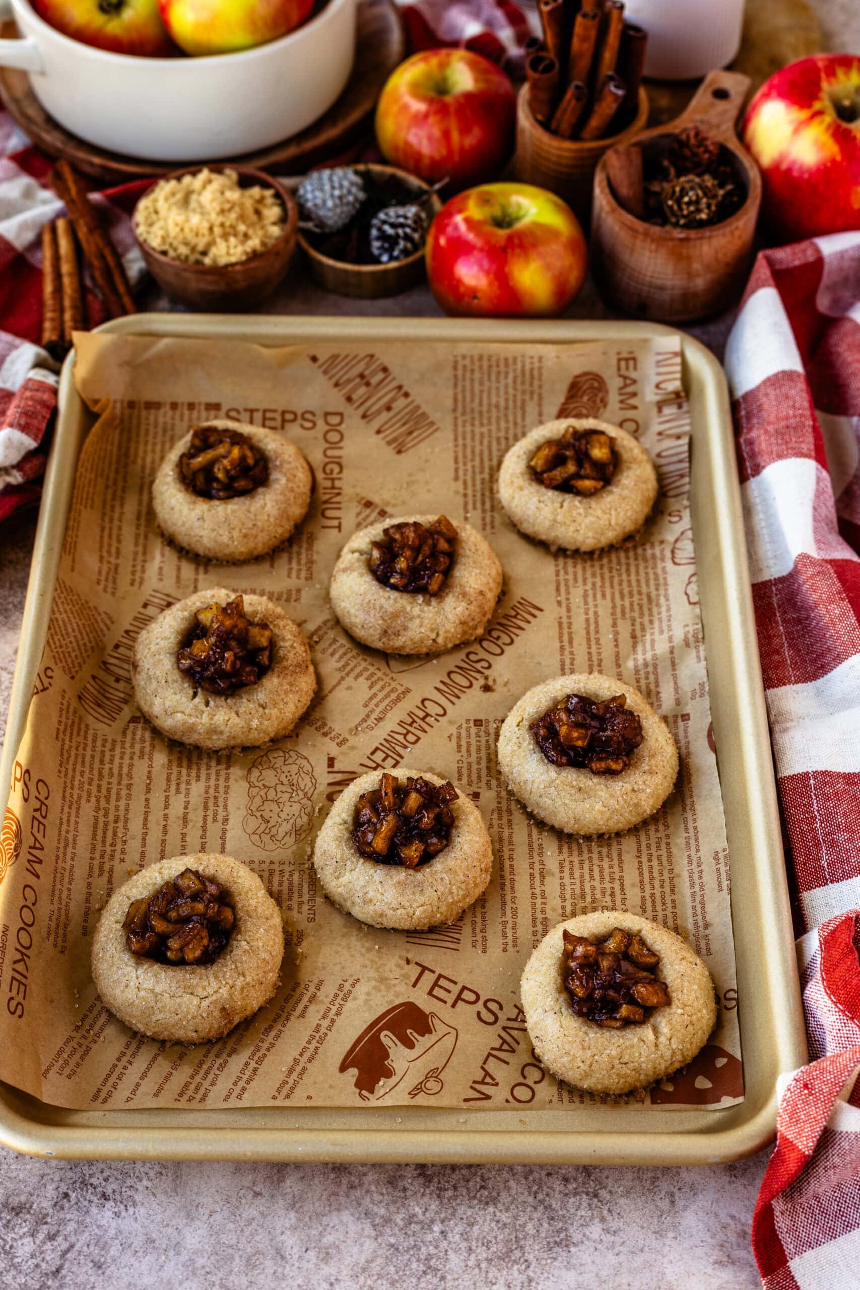 A baking tray filled with baked apple pie cookies, each with a cinnamon-sugar coating and a well of caramelized apple filling in the center. The cookies are arranged neatly on parchment paper printed with baking-themed illustrations. Fresh apples, brown sugar, cinnamon sticks, and pine cones are displayed in the background, creating a cozy and inviting autumn atmosphere.