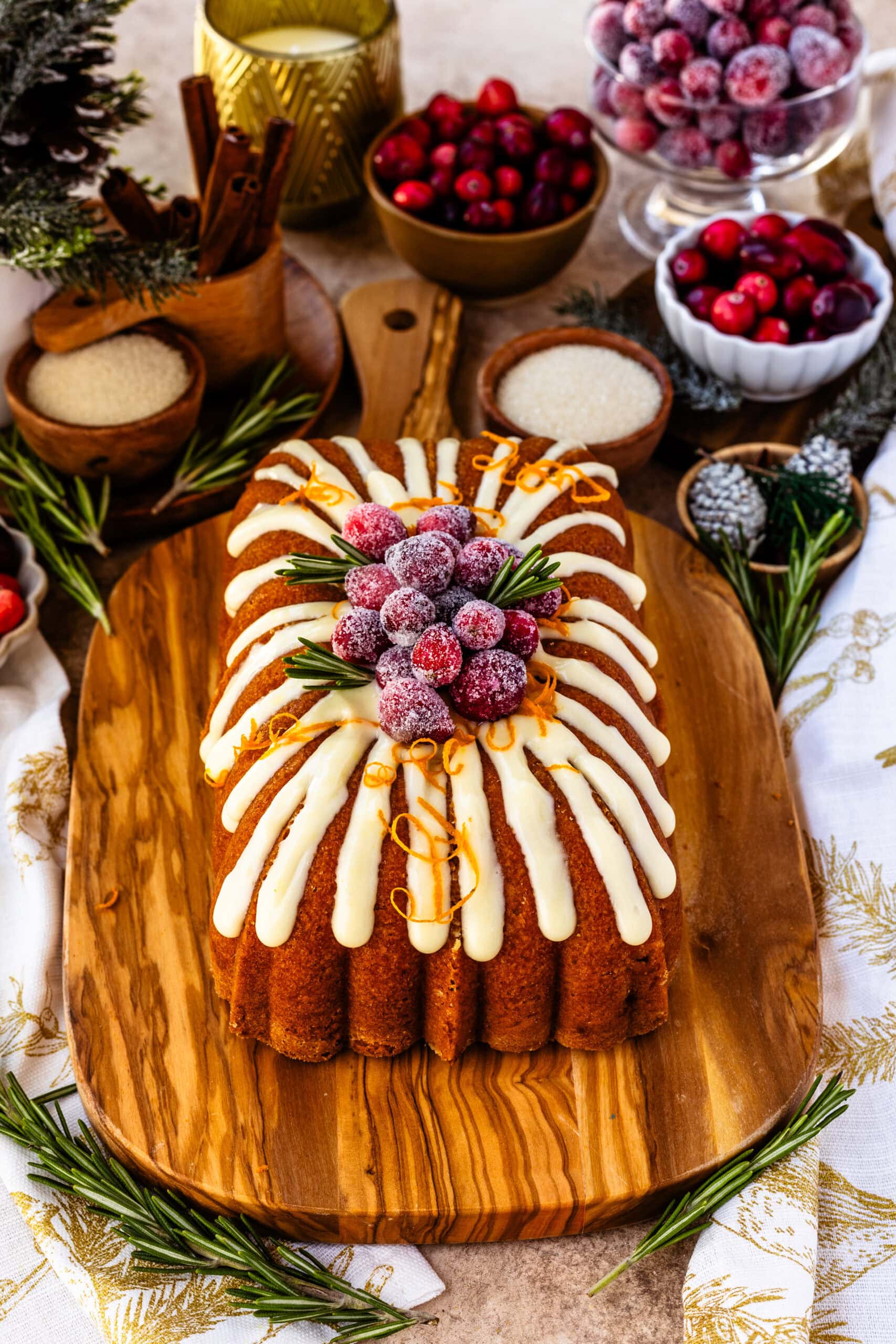 Orange cranberry bread topped with creamy orange frosting, sugared cranberries, fresh rosemary sprigs, and orange zest curls, beautifully presented on a wooden board. The festive garnishes and vibrant colors create an elegant holiday display, with the bread’s decorative design and cozy background enhancing the seasonal feel.