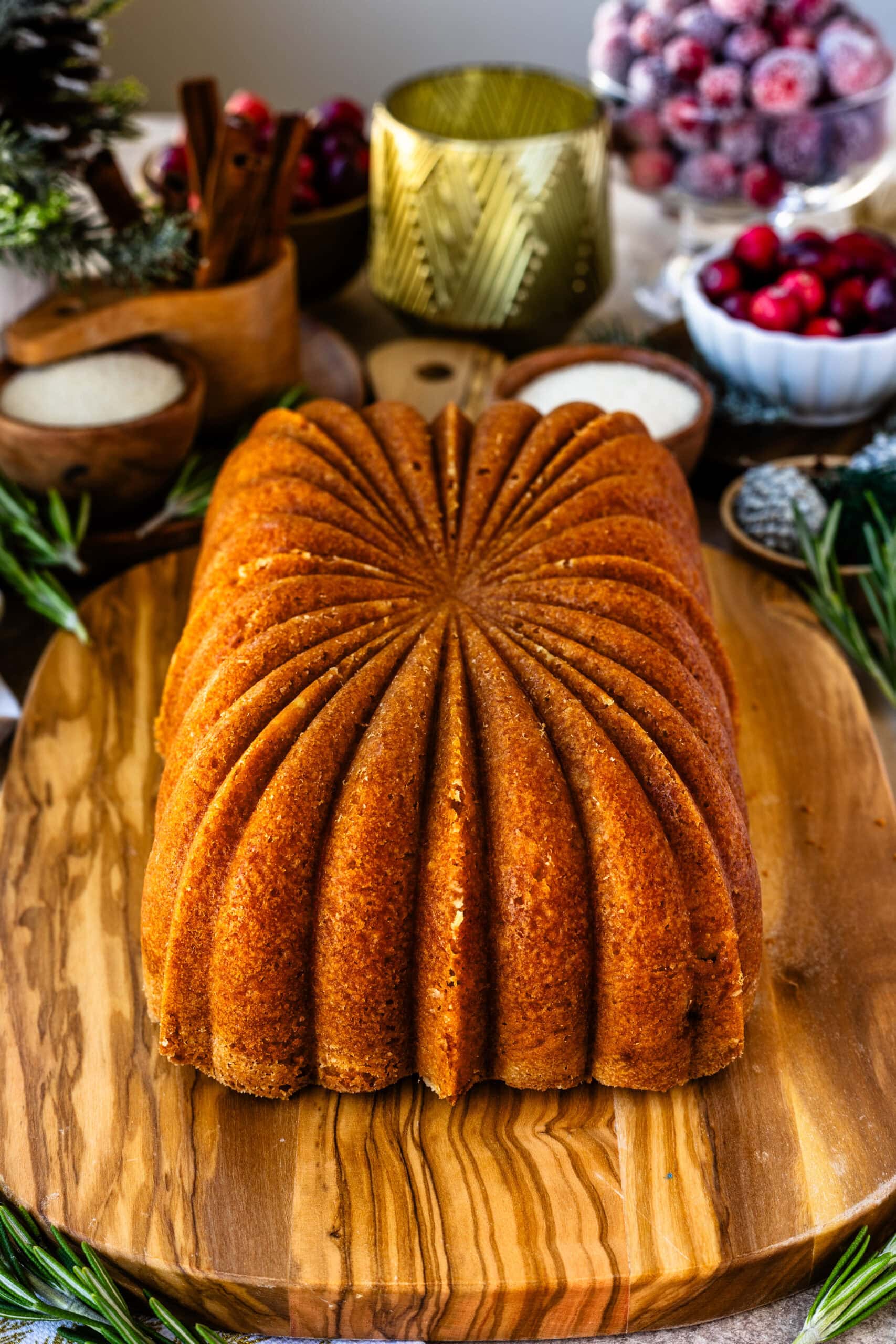Golden brown orange cranberry bread, freshly baked and displayed on a wooden board, showcasing a beautiful scalloped design on top. The background includes holiday decor elements like fresh cranberries, sugared cranberries, rosemary, and a candle, creating a festive and cozy atmosphere.