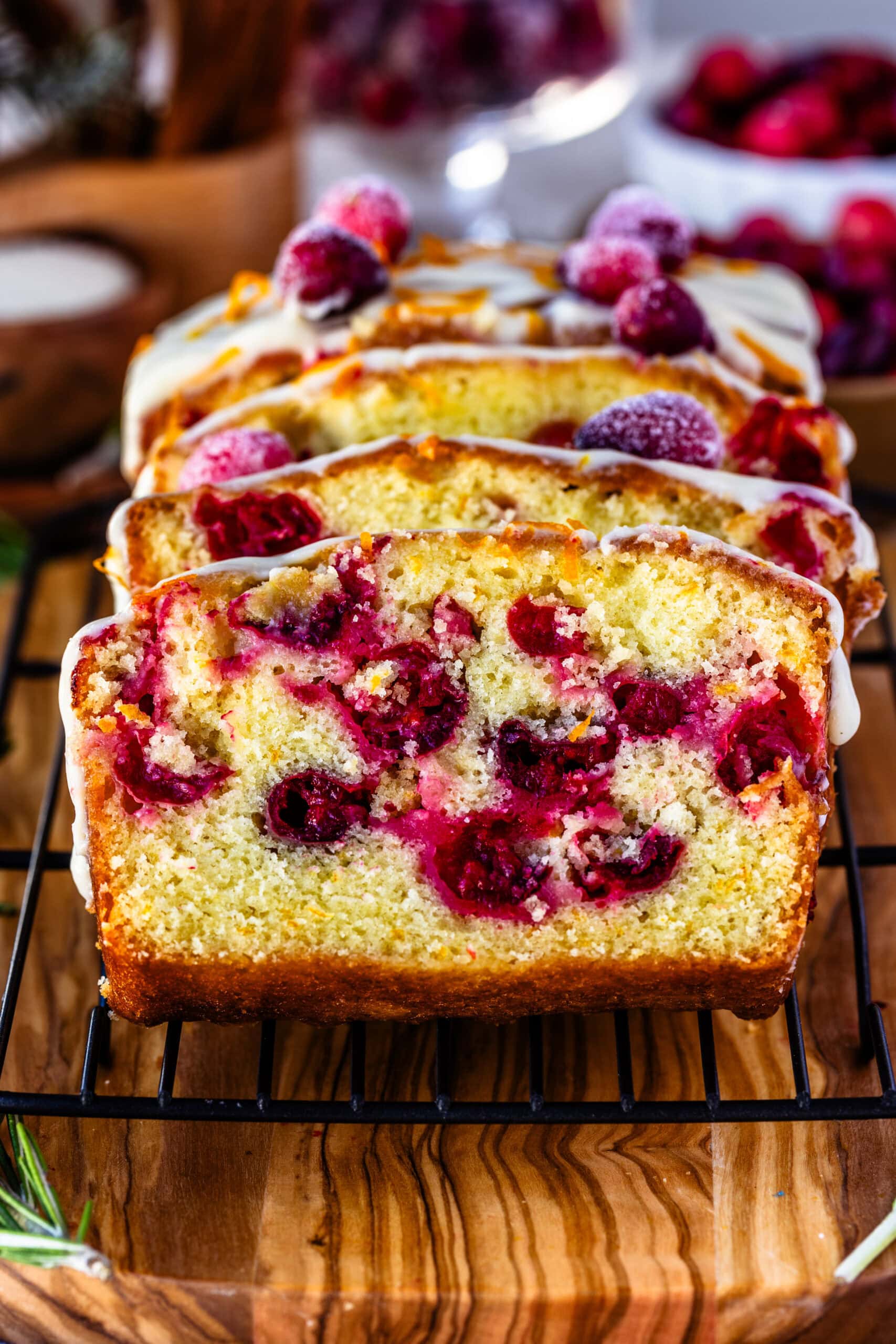 Close-up image of sliced orange cranberry bread on a cooling rack, topped with creamy orange frosting, fresh orange zest, and sugared cranberries. The vibrant cranberries are baked into the moist bread, creating a festive, colorful interior. The background features holiday elements like fresh cranberries and rosemary, adding a warm, seasonal feel to the presentation.