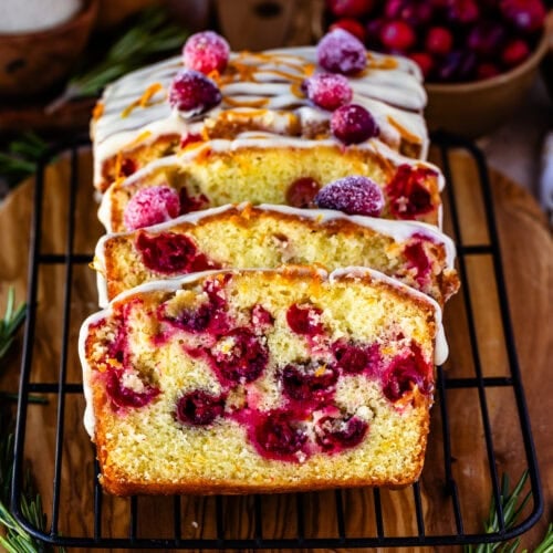 Close-up image of sliced orange cranberry bread on a cooling rack, topped with creamy orange frosting, fresh orange zest, and sugared cranberries. The vibrant cranberries are baked into the moist bread, creating a festive, colorful interior. The background features holiday elements like fresh cranberries and rosemary, adding a warm, seasonal feel to the presentation.