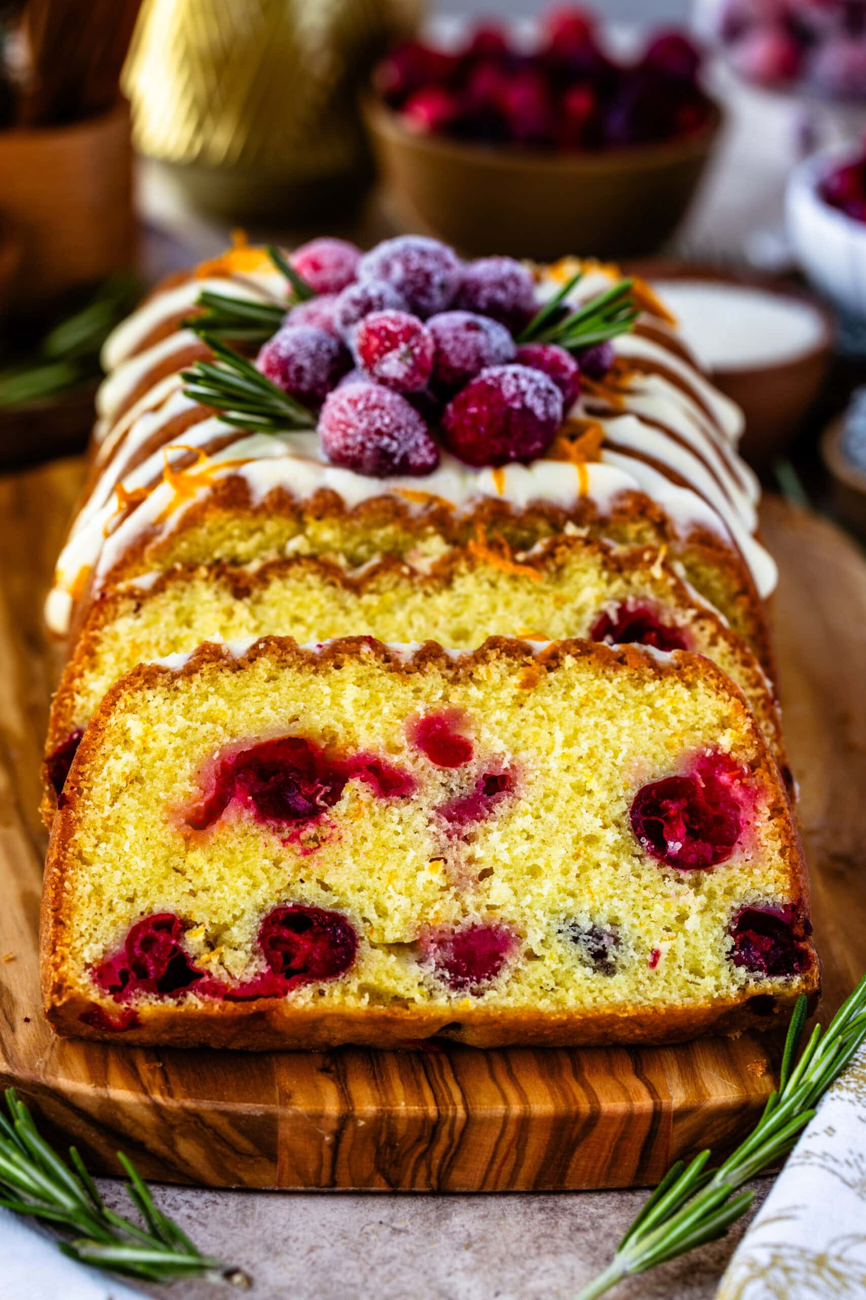 A close-up of a sliced orange cranberry bread loaf, revealing the moist, golden crumb studded with bright cranberries. The top is adorned with orange cream cheese frosting, sugared cranberries, fresh rosemary sprigs, and curls of orange zest, creating a festive presentation. Holiday elements, like cinnamon sticks and additional cranberries, add warmth and seasonal charm to the background.
