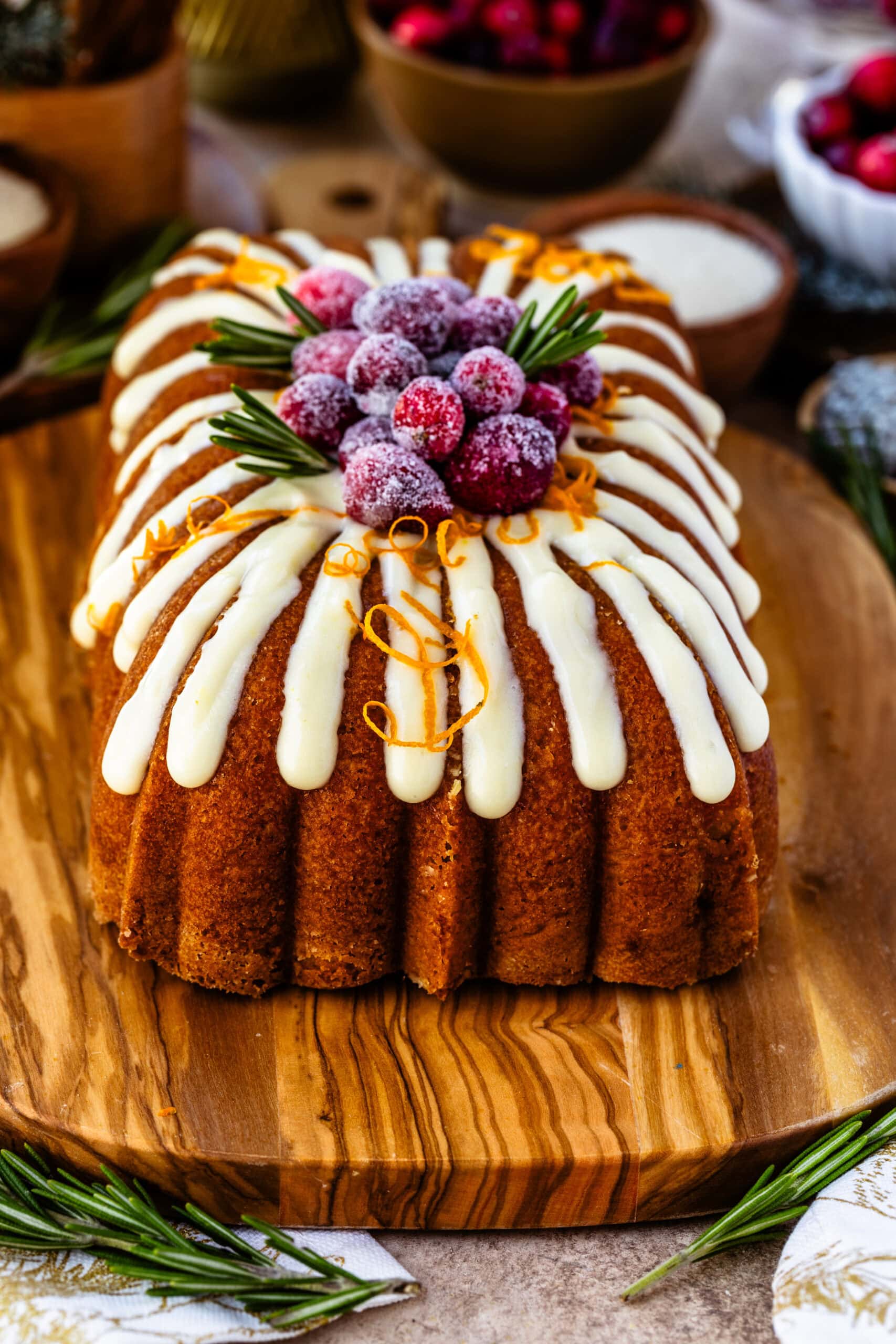 Orange cranberry bread topped with creamy orange frosting, sugared cranberries, fresh rosemary sprigs, and orange zest curls, beautifully presented on a wooden board. The festive garnishes and vibrant colors create an elegant holiday display, with the bread’s decorative design and cozy background enhancing the seasonal feel.