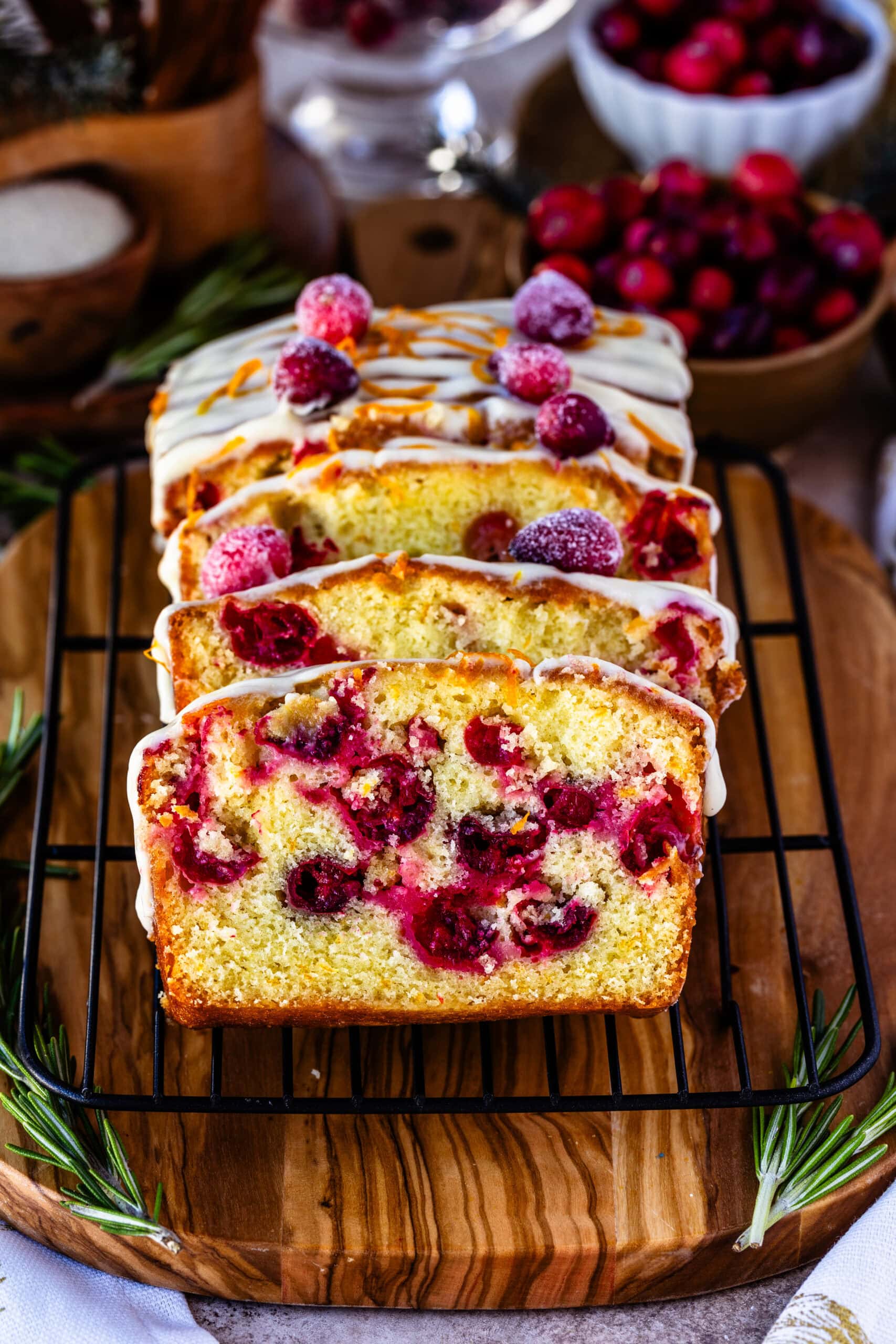 Close-up image of sliced orange cranberry bread on a cooling rack, topped with creamy orange frosting, fresh orange zest, and sugared cranberries. The vibrant cranberries are baked into the moist bread, creating a festive, colorful interior. The background features holiday elements like fresh cranberries and rosemary, adding a warm, seasonal feel to the presentation.