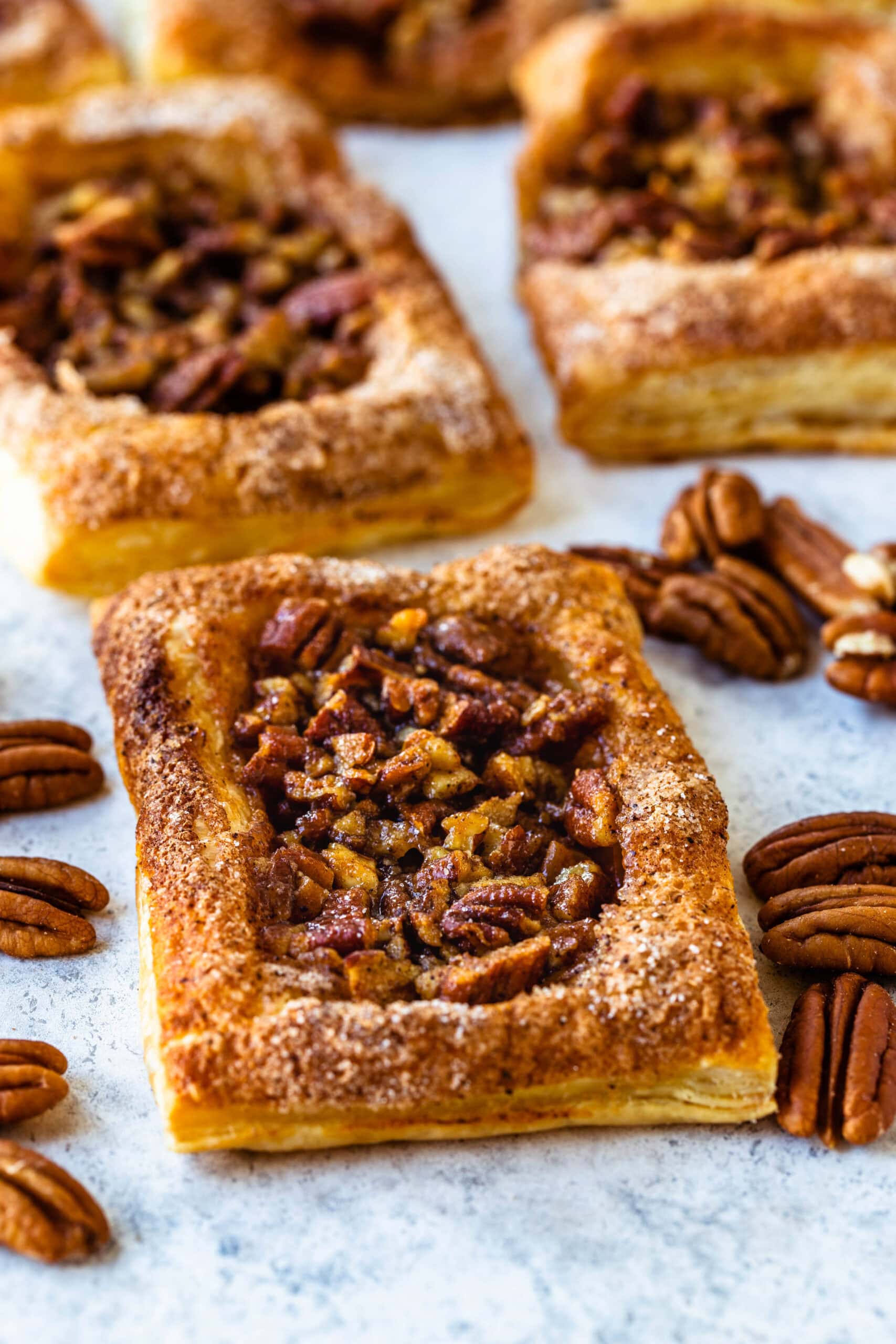 Close-up image of golden-brown Pecan Pie Puff Pastry squares, topped with crunchy pecans and surrounded by whole pecan halves on a light-colored background, showcasing the flaky, buttery texture of the pastry and the sweet, nutty filling.