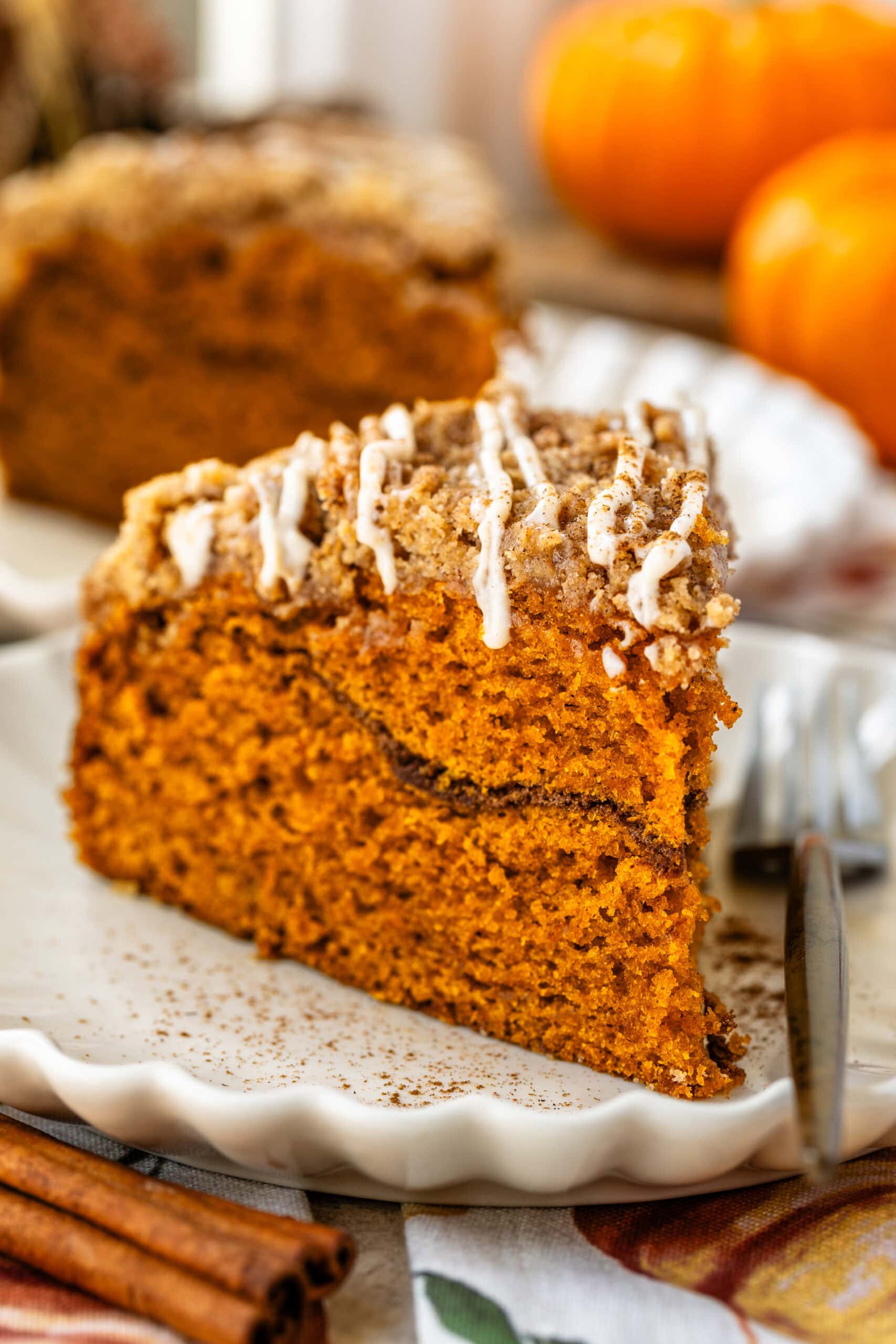 This image features a close-up of a perfectly sliced piece of pumpkin coffee cake, showcasing its moist and fluffy interior with a visible cinnamon sugar layer in the middle. The crumbly streusel topping is drizzled with a glaze, adding extra sweetness and texture. The cake is plated beautifully on a scalloped-edge plate, and the warm, cozy atmosphere with pumpkins in the background enhances the seasonal autumn feel of this dessert.