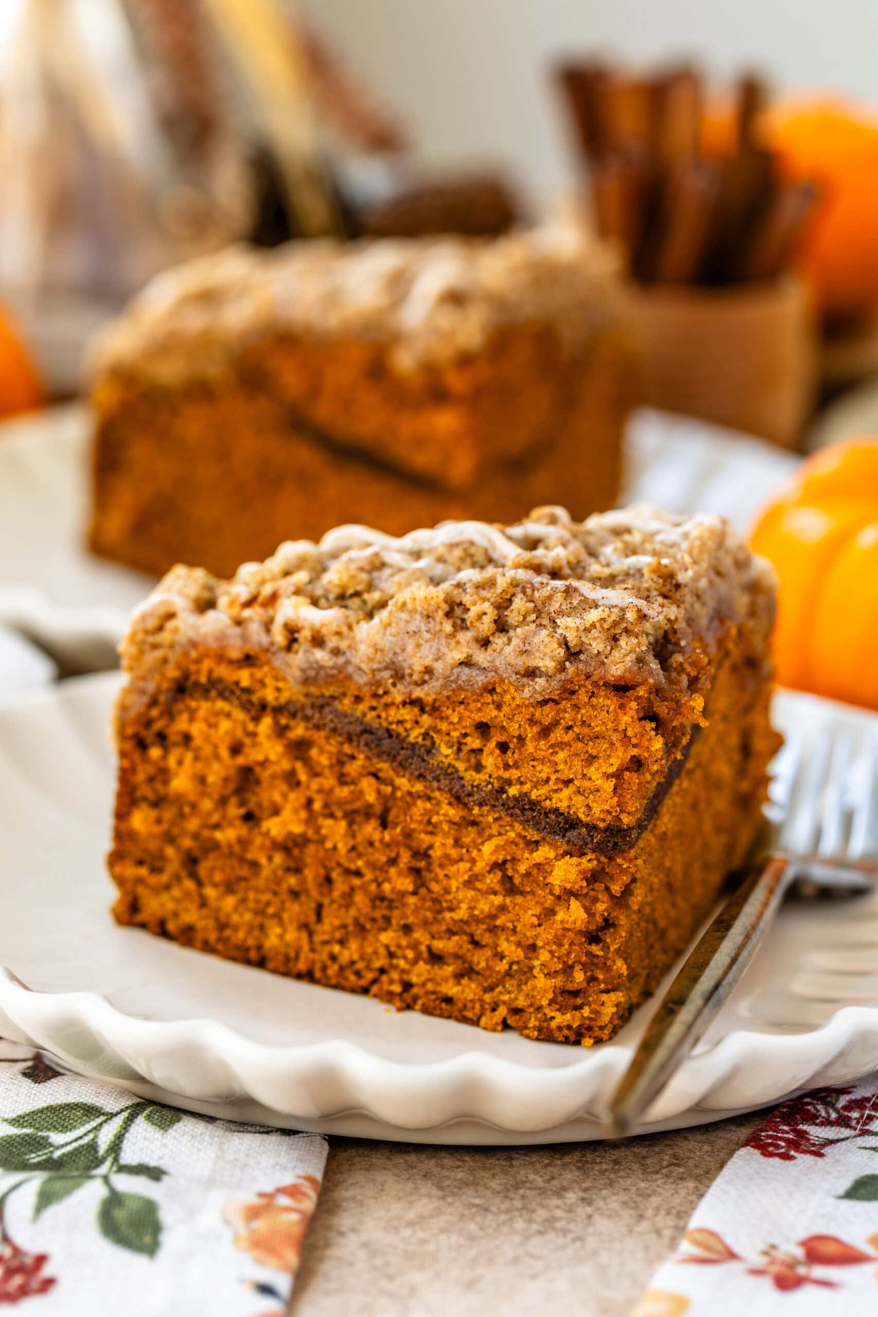 This image features a close-up of a perfectly sliced piece of pumpkin coffee cake, showcasing its moist and fluffy interior with a visible cinnamon sugar layer in the middle. The crumbly streusel topping is drizzled with a glaze, adding extra sweetness and texture. The cake is plated beautifully on a scalloped-edge plate, and the warm, cozy atmosphere with pumpkins in the background enhances the seasonal autumn feel of this dessert.