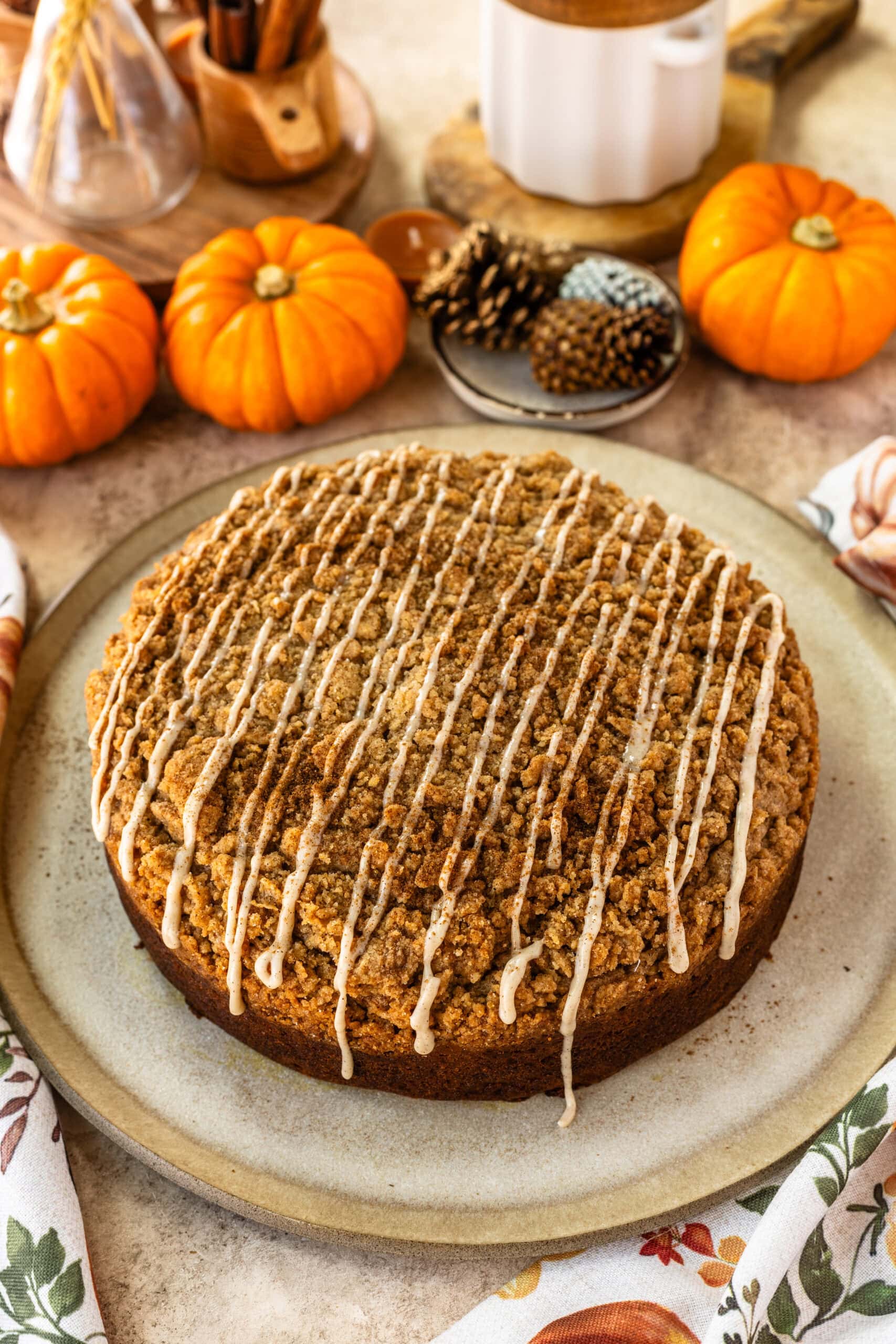 This image showcases a finished pumpkin coffee cake, topped with a generous drizzle of creamy glaze over its streusel topping. The cake sits on a rustic plate, with decorative pumpkins, pinecones, and cinnamon sticks in the background, evoking a cozy fall atmosphere. The zigzag pattern of the glaze adds a beautiful touch, making the cake look both festive and delicious.
