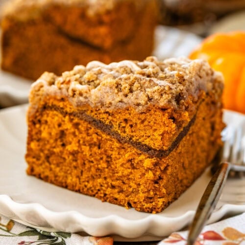 This image features a close-up of a perfectly sliced piece of pumpkin coffee cake, showcasing its moist and fluffy interior with a visible cinnamon sugar layer in the middle. The crumbly streusel topping is drizzled with a glaze, adding extra sweetness and texture. The cake is plated beautifully on a scalloped-edge plate, and the warm, cozy atmosphere with pumpkins in the background enhances the seasonal autumn feel of this dessert.