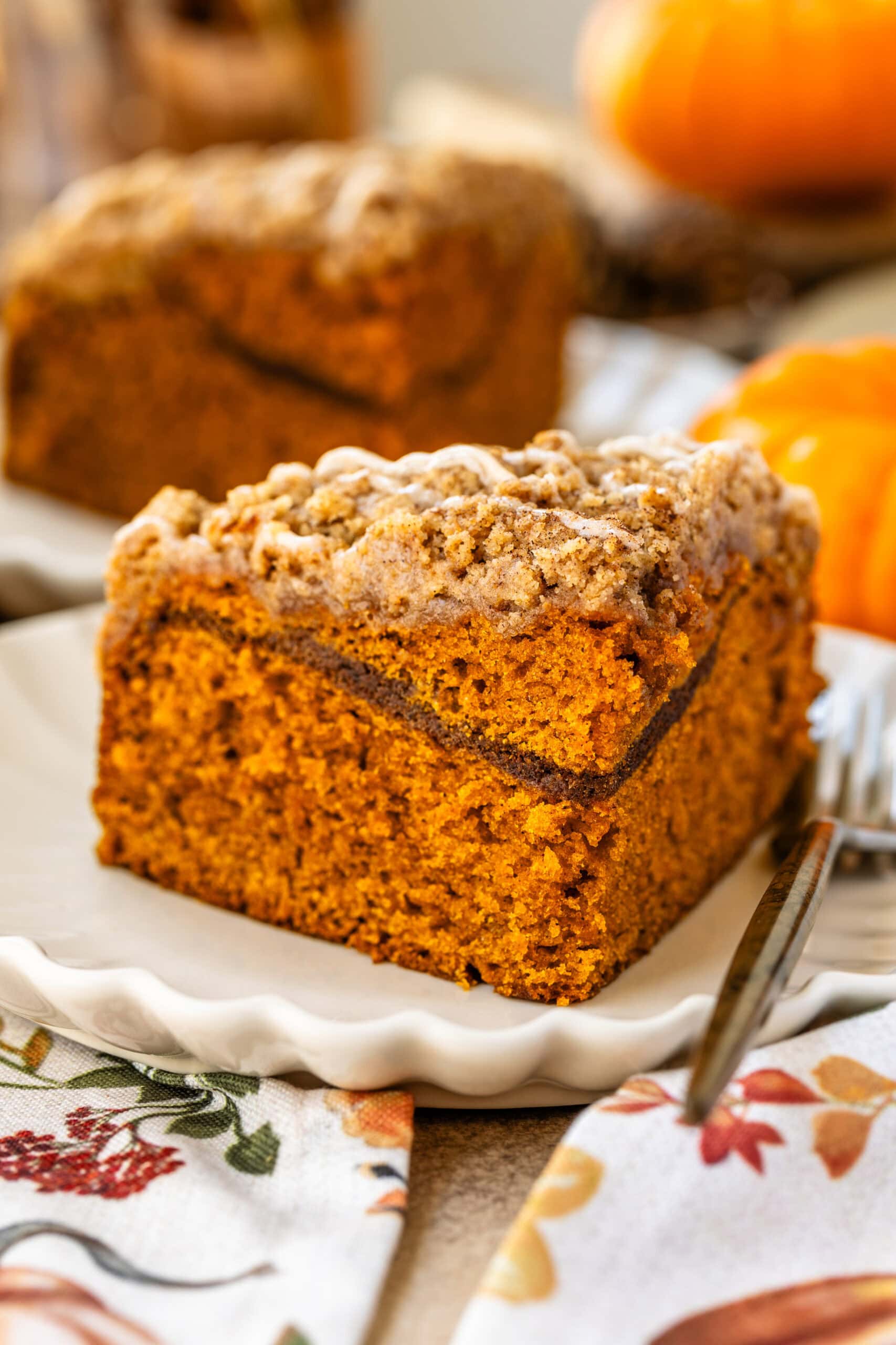 This image features a close-up of a perfectly sliced piece of pumpkin coffee cake, showcasing its moist and fluffy interior with a visible cinnamon sugar layer in the middle. The crumbly streusel topping is drizzled with a glaze, adding extra sweetness and texture. The cake is plated beautifully on a scalloped-edge plate, and the warm, cozy atmosphere with pumpkins in the background enhances the seasonal autumn feel of this dessert.