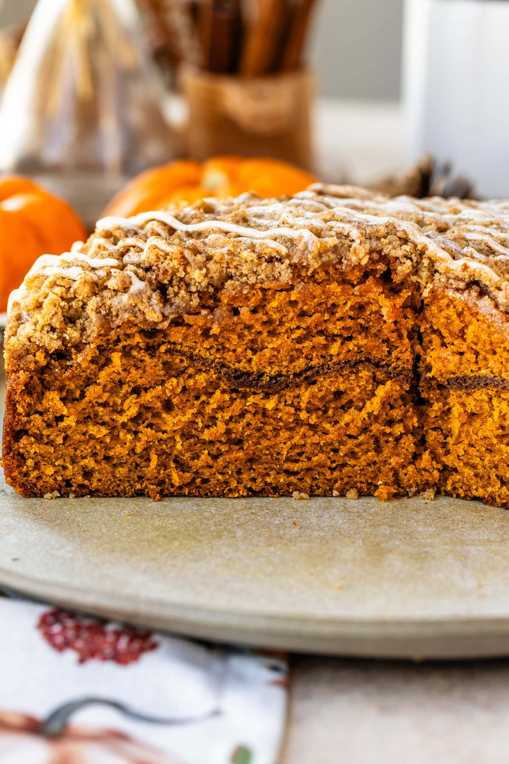This image displays a cross-section of a beautifully baked pumpkin coffee cake, revealing its moist, tender interior with a layer of cinnamon sugar in the middle. The streusel topping is finished with a drizzle of glaze, adding a sweet touch to the crunchy top. The warm, orange hue of the cake is complemented by the cozy autumn setting with pumpkins and pinecones in the background, highlighting the seasonal appeal of this dessert.