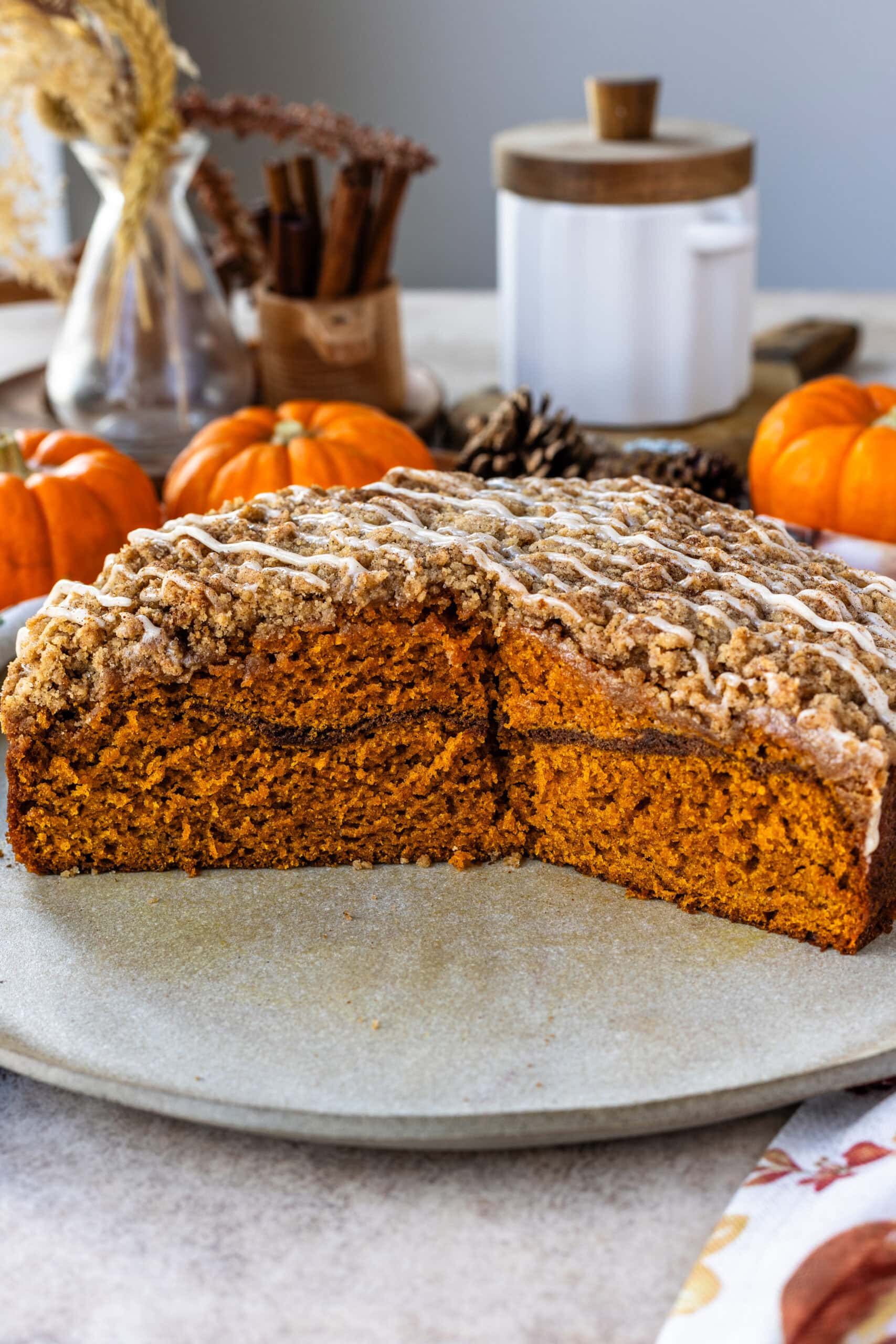 This image displays a cross-section of a beautifully baked pumpkin coffee cake, revealing its moist, tender interior with a layer of cinnamon sugar in the middle. The streusel topping is finished with a drizzle of glaze, adding a sweet touch to the crunchy top. The warm, orange hue of the cake is complemented by the cozy autumn setting with pumpkins and pinecones in the background, highlighting the seasonal appeal of this dessert.