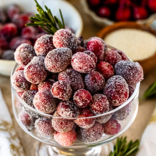 This image shows a beautiful close-up of sugared cranberries in a clear glass bowl. Each cranberry is coated with a layer of sugar, giving them a frosty, festive appearance. The cranberries are paired with a sprig of rosemary, adding a touch of green for a holiday vibe. The background features blurred cranberries and holiday-themed decor, enhancing the seasonal atmosphere. This display of sugared cranberries is perfect for a holiday table or as a dessert garnish.