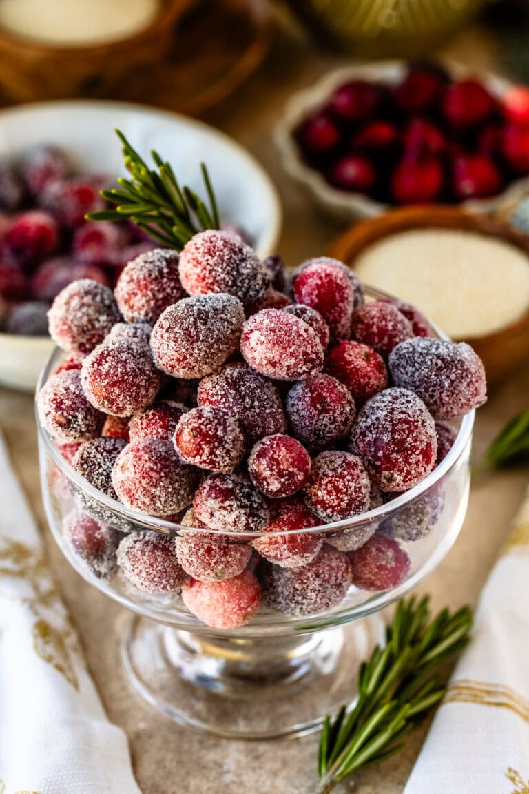 This image shows a beautiful close-up of sugared cranberries in a clear glass bowl. Each cranberry is coated with a layer of sugar, giving them a frosty, festive appearance. The cranberries are paired with a sprig of rosemary, adding a touch of green for a holiday vibe. The background features blurred cranberries and holiday-themed decor, enhancing the seasonal atmosphere. This display of sugared cranberries is perfect for a holiday table or as a dessert garnish.