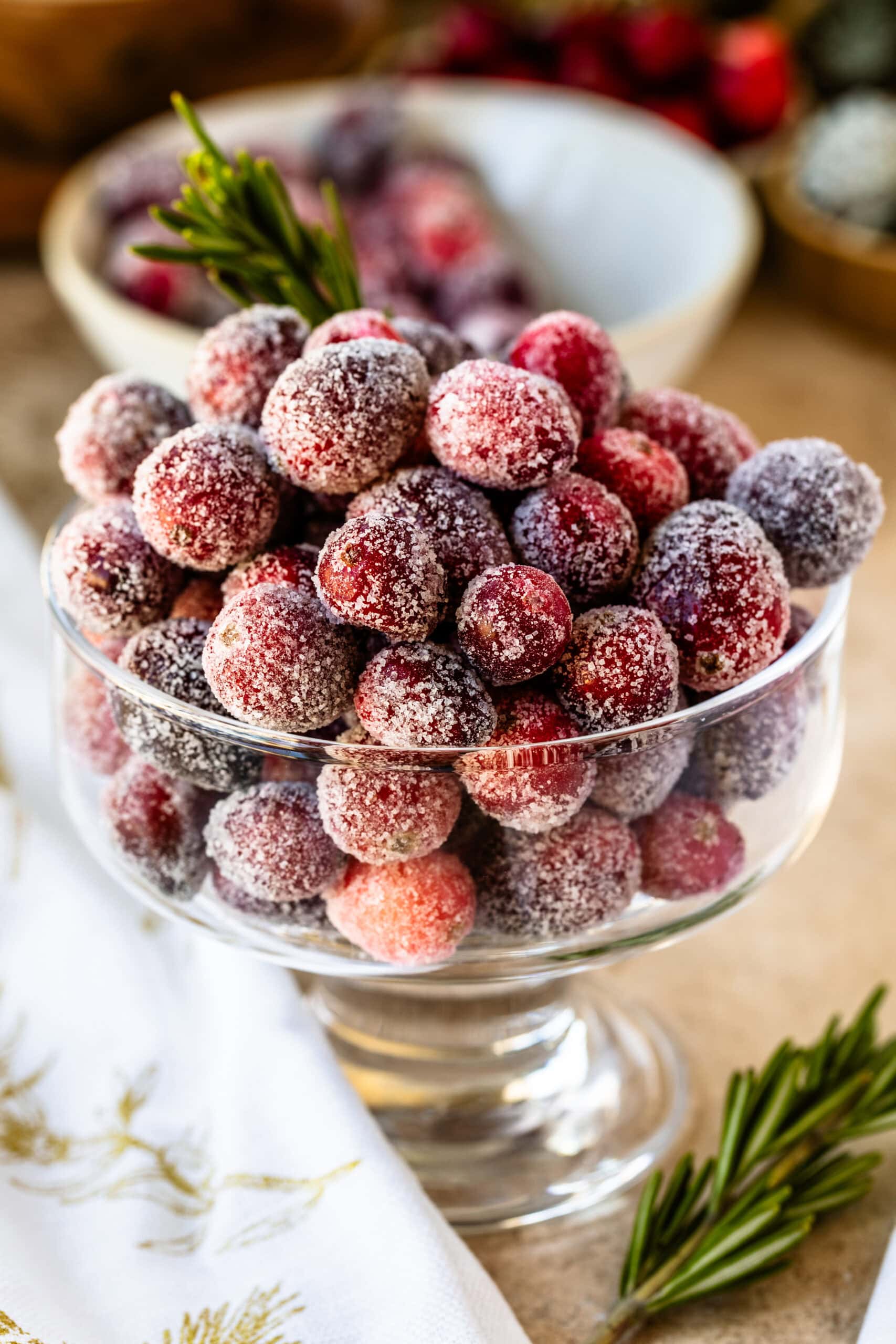 This image shows a beautiful close-up of sugared cranberries in a clear glass bowl. Each cranberry is coated with a layer of sugar, giving them a frosty, festive appearance. The cranberries are paired with a sprig of rosemary, adding a touch of green for a holiday vibe. The background features blurred cranberries and holiday-themed decor, enhancing the seasonal atmosphere. This display of sugared cranberries is perfect for a holiday table or as a dessert garnish.