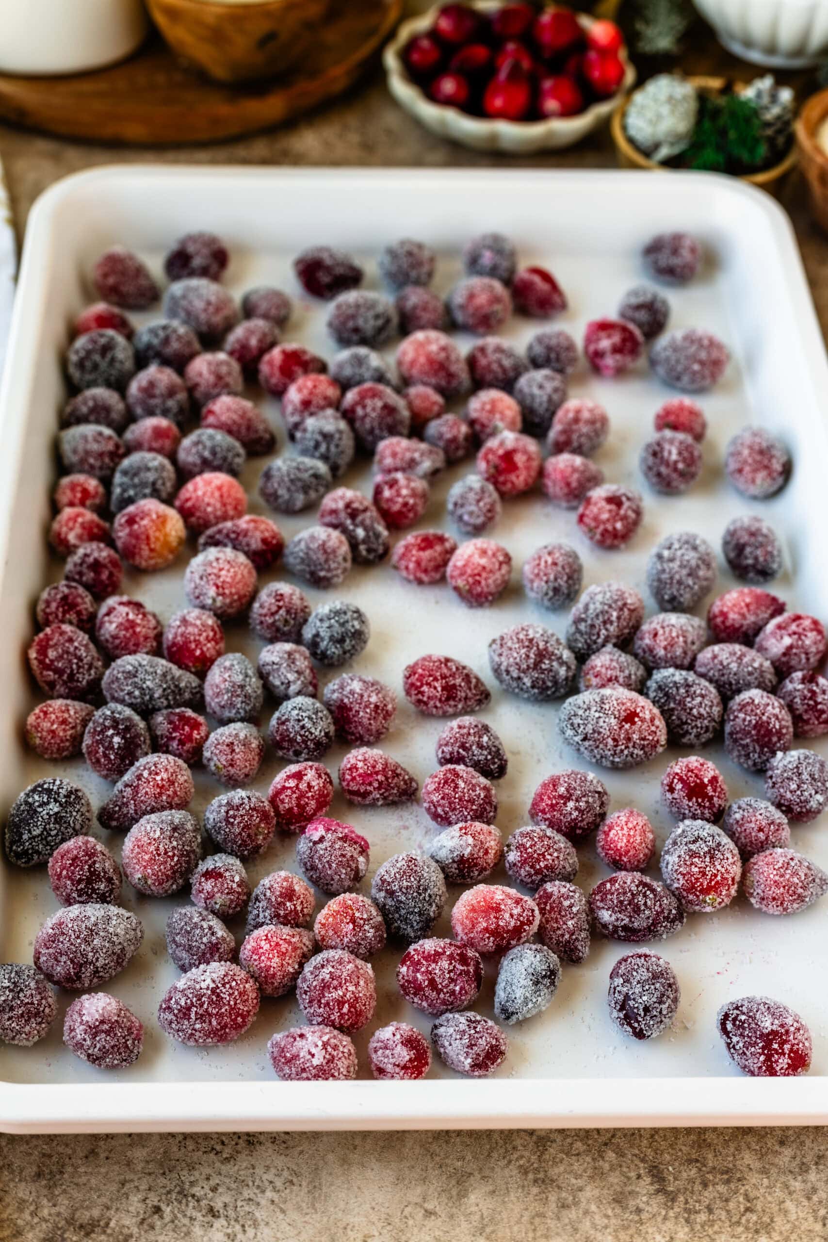 This image shows a tray of sugared cranberries spread out on a white baking sheet, allowing them to dry evenly. The cranberries are coated in a layer of sugar, giving them a frosted, crystalline appearance. In the background, you can see additional holiday elements such as bowls of cranberries and rustic decorations, creating a festive and cozy atmosphere. The sugared cranberries are ready to be used as garnishes or holiday snacks.