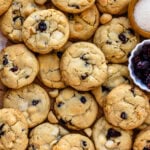 Overhead view of a batch of white chocolate cranberry cookies arranged closely together, highlighting their golden-brown color with visible pieces of white chocolate and dried cranberries. Surrounding the cookies are small bowls of macadamia nuts, sugar, and white chocolate chips, adding visual interest and showcasing the ingredients in the recipe.