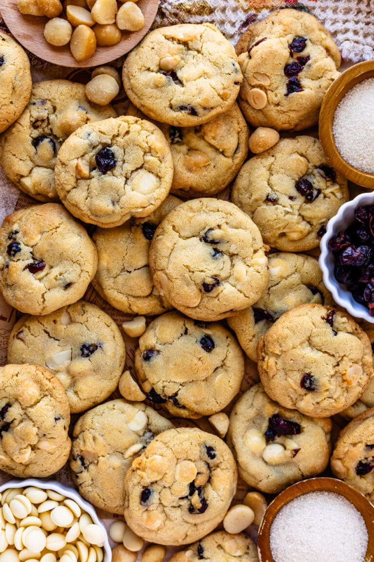 Overhead view of a batch of white chocolate cranberry cookies arranged closely together, highlighting their golden-brown color with visible pieces of white chocolate and dried cranberries. Surrounding the cookies are small bowls of macadamia nuts, sugar, and white chocolate chips, adding visual interest and showcasing the ingredients in the recipe.