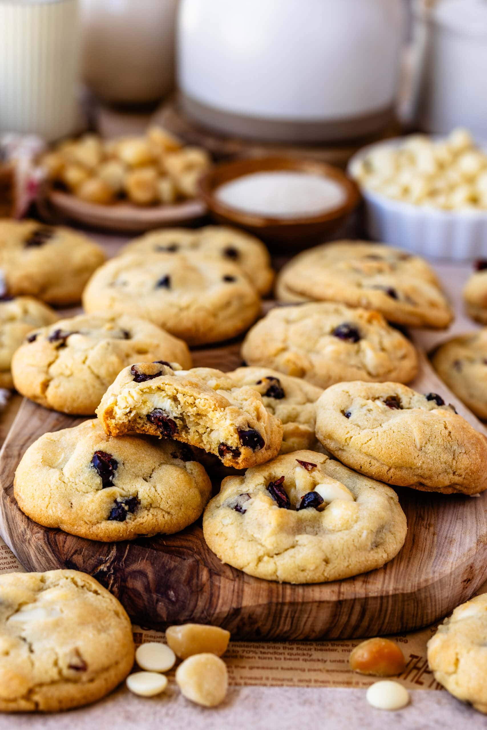 Close-up of a stack of white chocolate cranberry cookies with the top cookie broken in half, showing a soft, chewy interior with chunks of white chocolate and dried cranberries. The golden-brown edges contrast with the soft center, emphasizing the cookie's texture. A few macadamia nut pieces are scattered around, adding to the inviting presentation.
