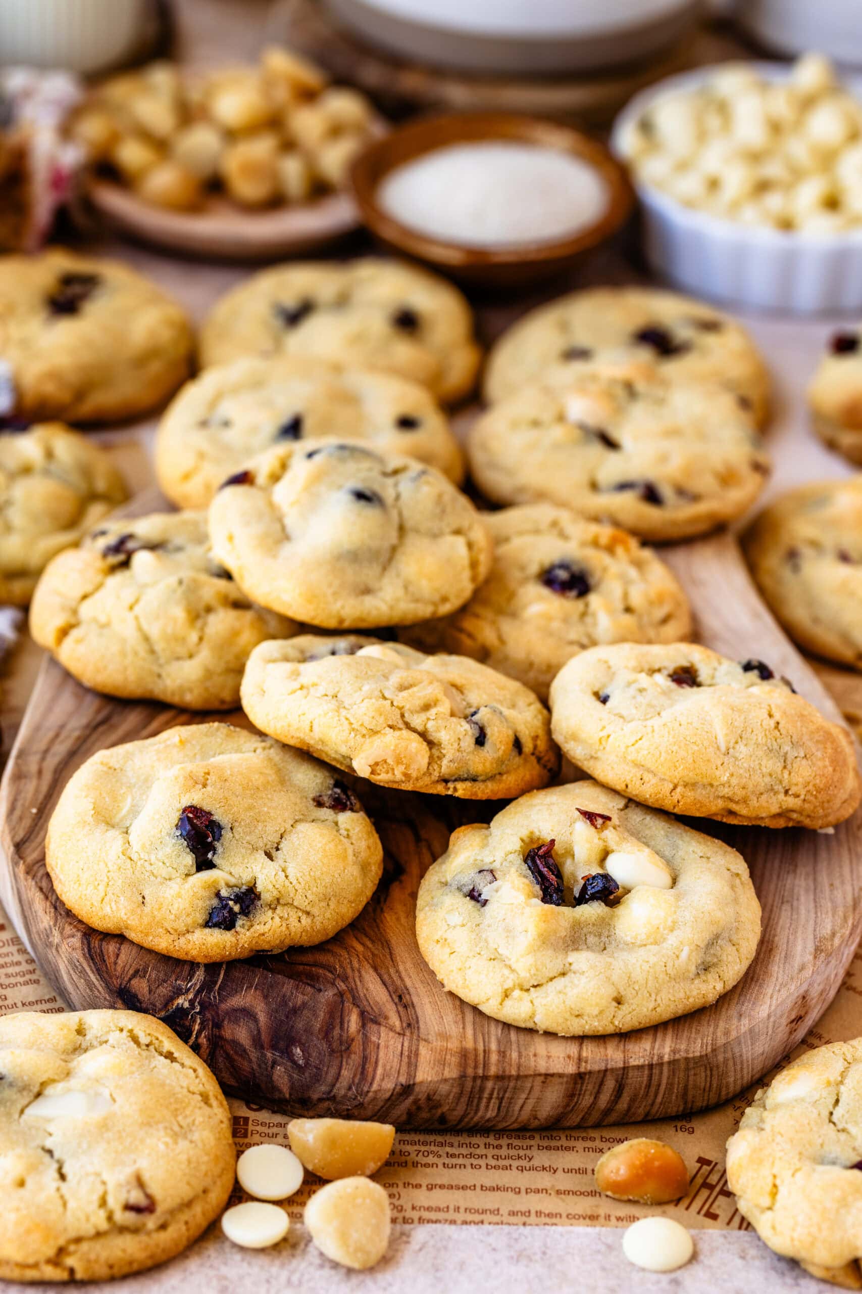 A plate of freshly baked cookies stacked on a round serving tray. The cookies are golden brown, with visible chunks of white chocolate and dried cranberries throughout. The background includes small bowls with ingredients like macadamia nuts, sugar, and white chocolate chips, creating a warm, inviting atmosphere that highlights the cookie ingredients.
