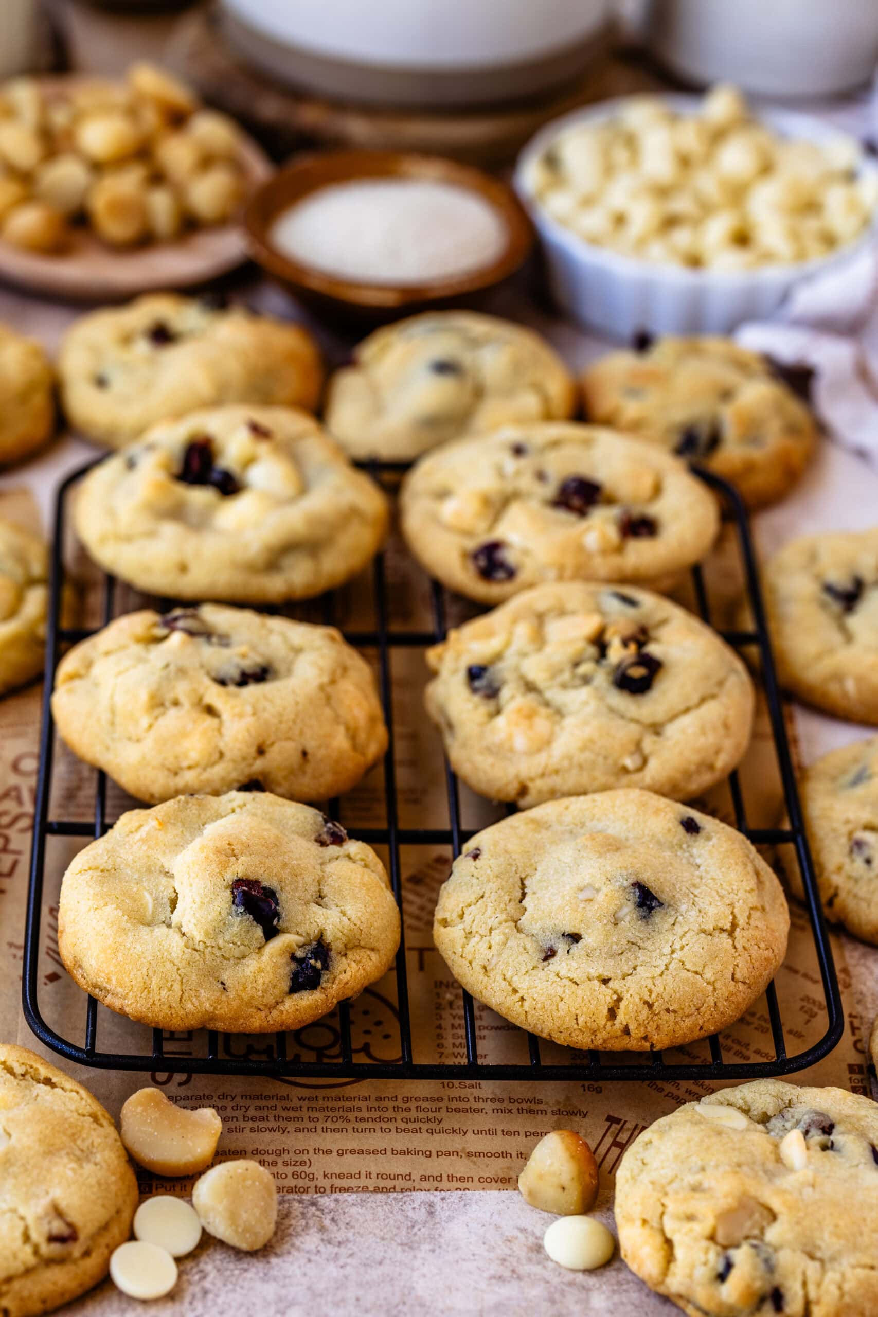 White chocolate cranberry cookies cooling on a wire rack, showcasing their golden-brown color with visible chunks of white chocolate and dried cranberries. The background includes small bowls filled with macadamia nuts, sugar, and white chocolate chips, adding to the cozy, homemade baking atmosphere.