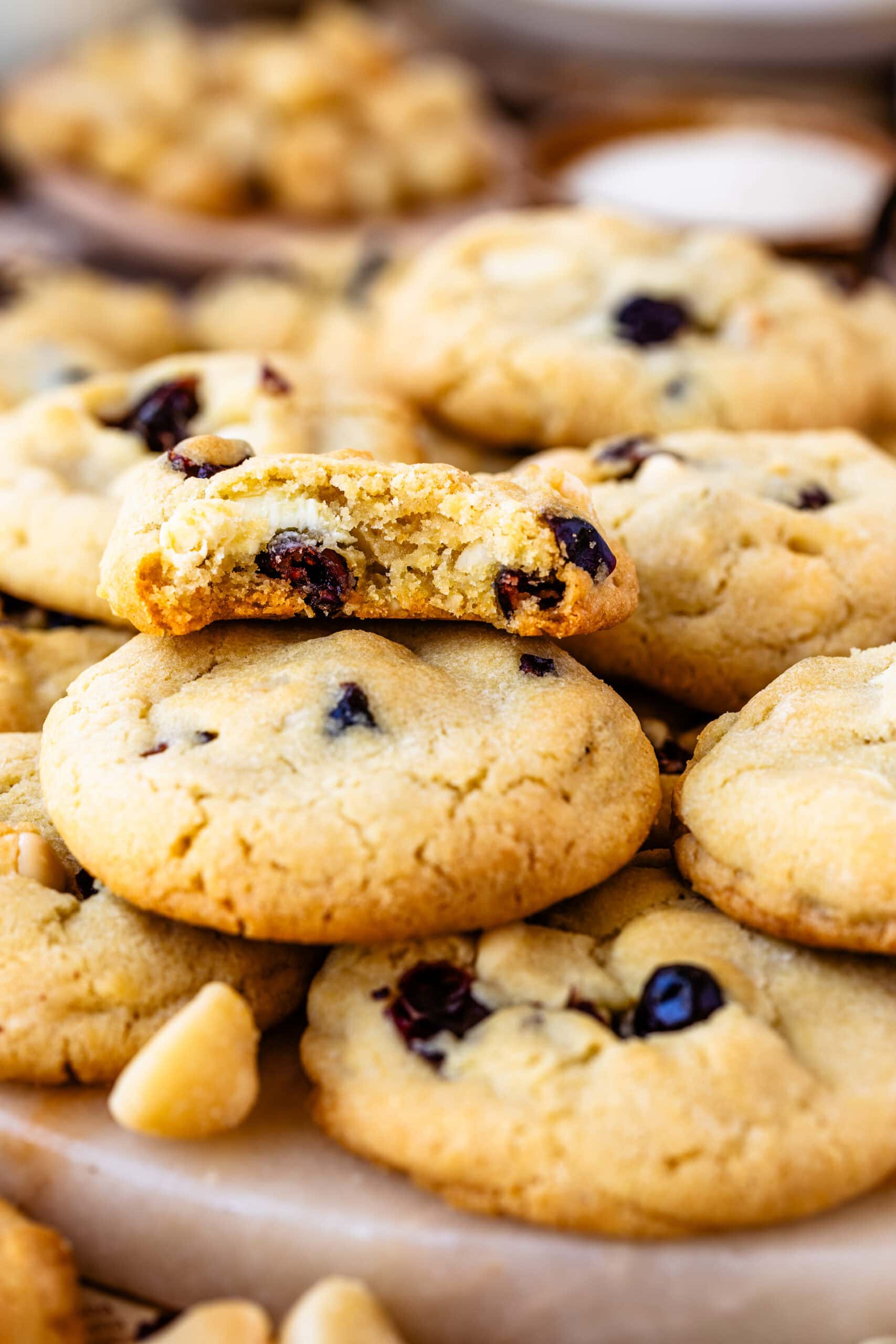 Close-up of a stack of white chocolate cranberry cookies with the top cookie broken in half, showing a soft, chewy interior with chunks of white chocolate and dried cranberries. The golden-brown edges contrast with the soft center, emphasizing the cookie's texture. A few macadamia nut pieces are scattered around, adding to the inviting presentation.