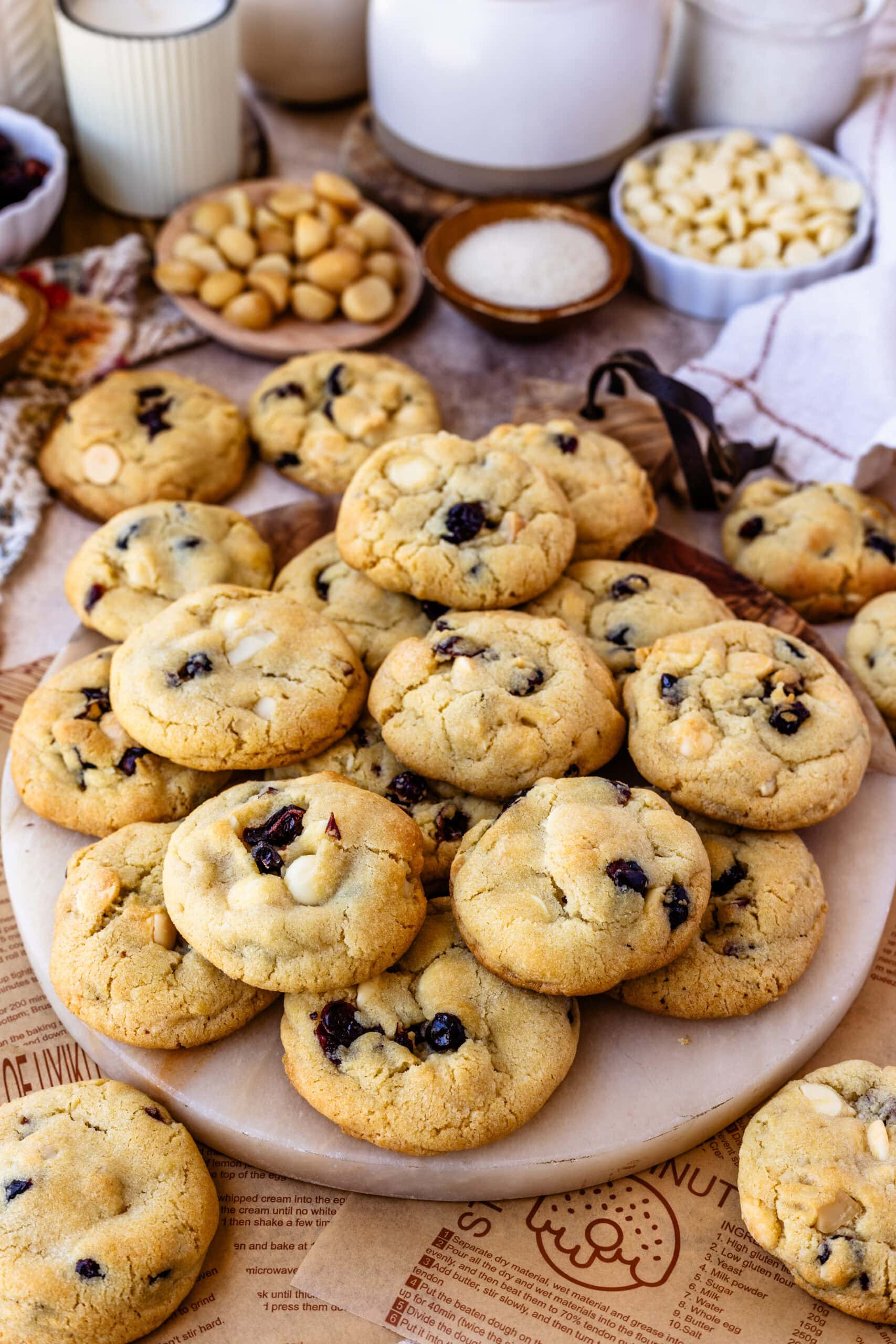 A plate of freshly baked cookies stacked on a round serving tray. The cookies are golden brown, with visible chunks of white chocolate and dried cranberries throughout. The background includes small bowls with ingredients like macadamia nuts, sugar, and white chocolate chips, creating a warm, inviting atmosphere that highlights the cookie ingredients.