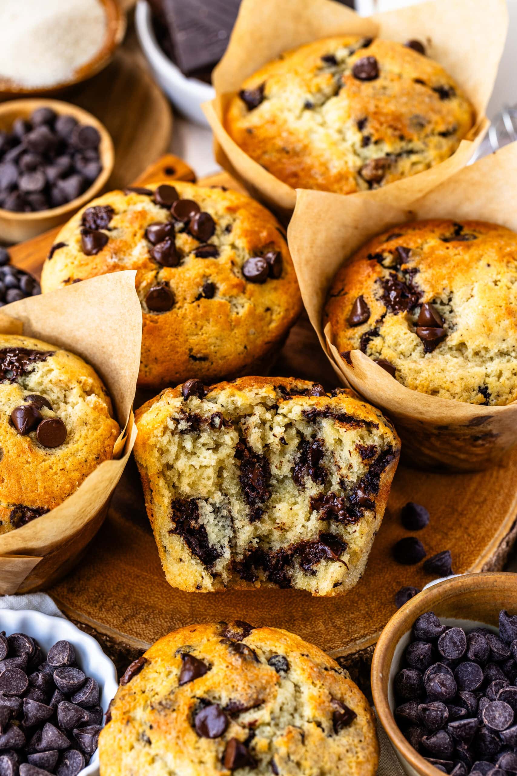 The image shows a close-up of several chocolate chip muffins, with one muffin cut in half to reveal its soft, fluffy interior filled with gooey melted chocolate chips. The muffins are perfectly golden on the outside, while the inside looks moist and tender. Surrounding the muffins are more chocolate chips in small bowls, adding to the indulgent and inviting atmosphere. The tulip-style parchment paper liners enhance the homemade, rustic appeal of these delicious treats.