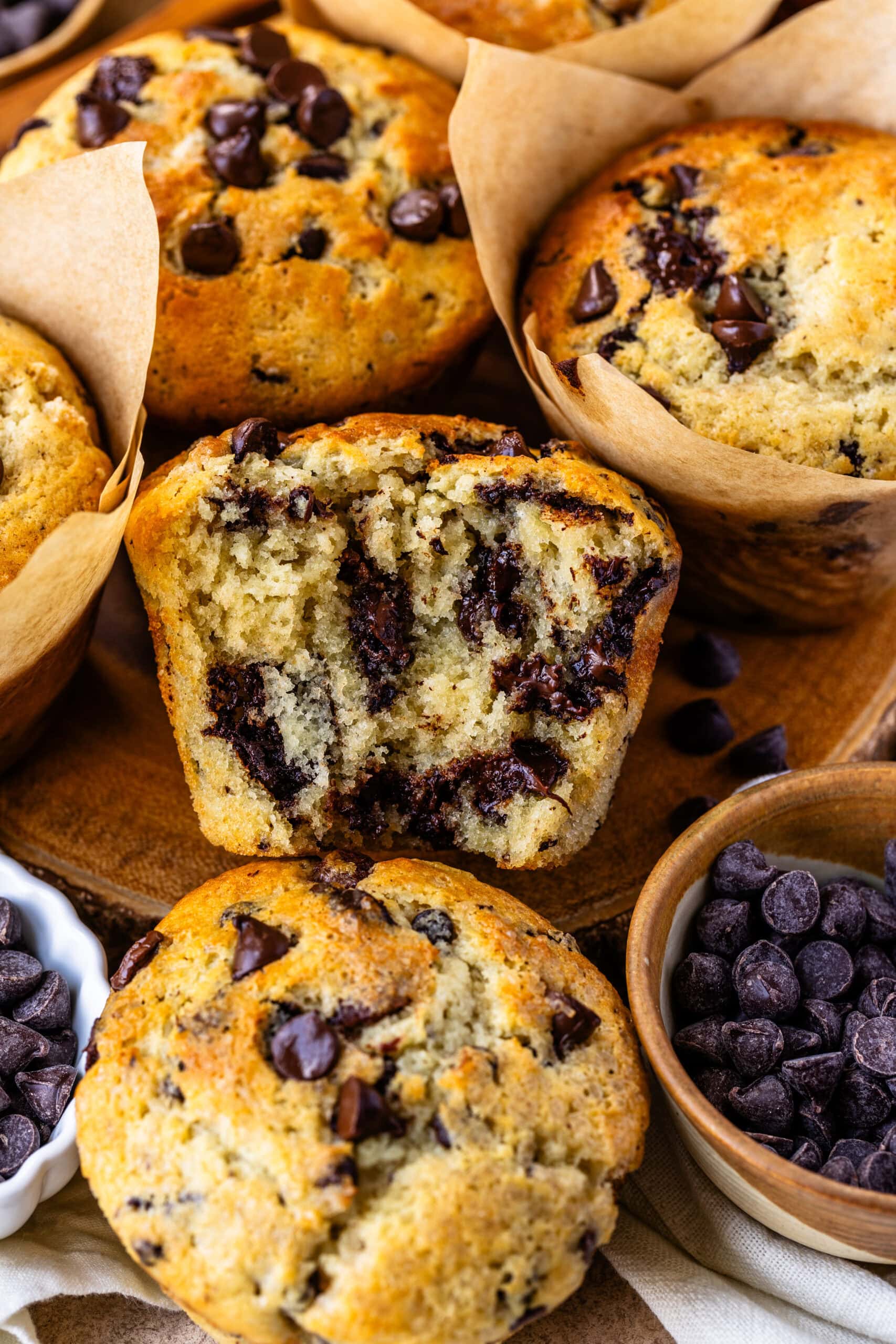 The image shows a close-up of several chocolate chip muffins, with one muffin cut in half to reveal its soft, fluffy interior filled with gooey melted chocolate chips. The muffins are perfectly golden on the outside, while the inside looks moist and tender. Surrounding the muffins are more chocolate chips in small bowls, adding to the indulgent and inviting atmosphere. The tulip-style parchment paper liners enhance the homemade, rustic appeal of these delicious treats.