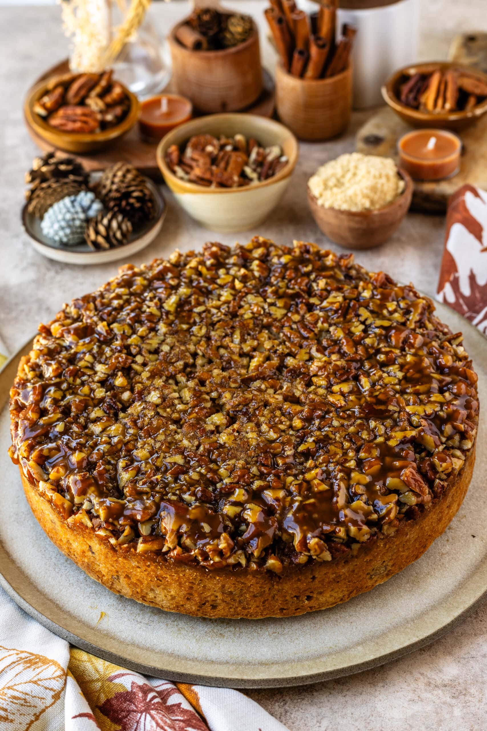 The image shows a beautifully baked pecan pie cake displayed on a neutral-colored plate. The cake is topped with a generous layer of caramelized, chopped pecans, glistening with a rich caramel drizzle that adds a decadent finish. The warm, golden-brown tones of the cake are complemented by the cozy autumnal background, which features bowls of pecans, brown sugar, cinnamon sticks, and candles. The surrounding decor enhances the inviting and seasonal feel, making the cake look perfect for a fall gathering or holiday dessert.