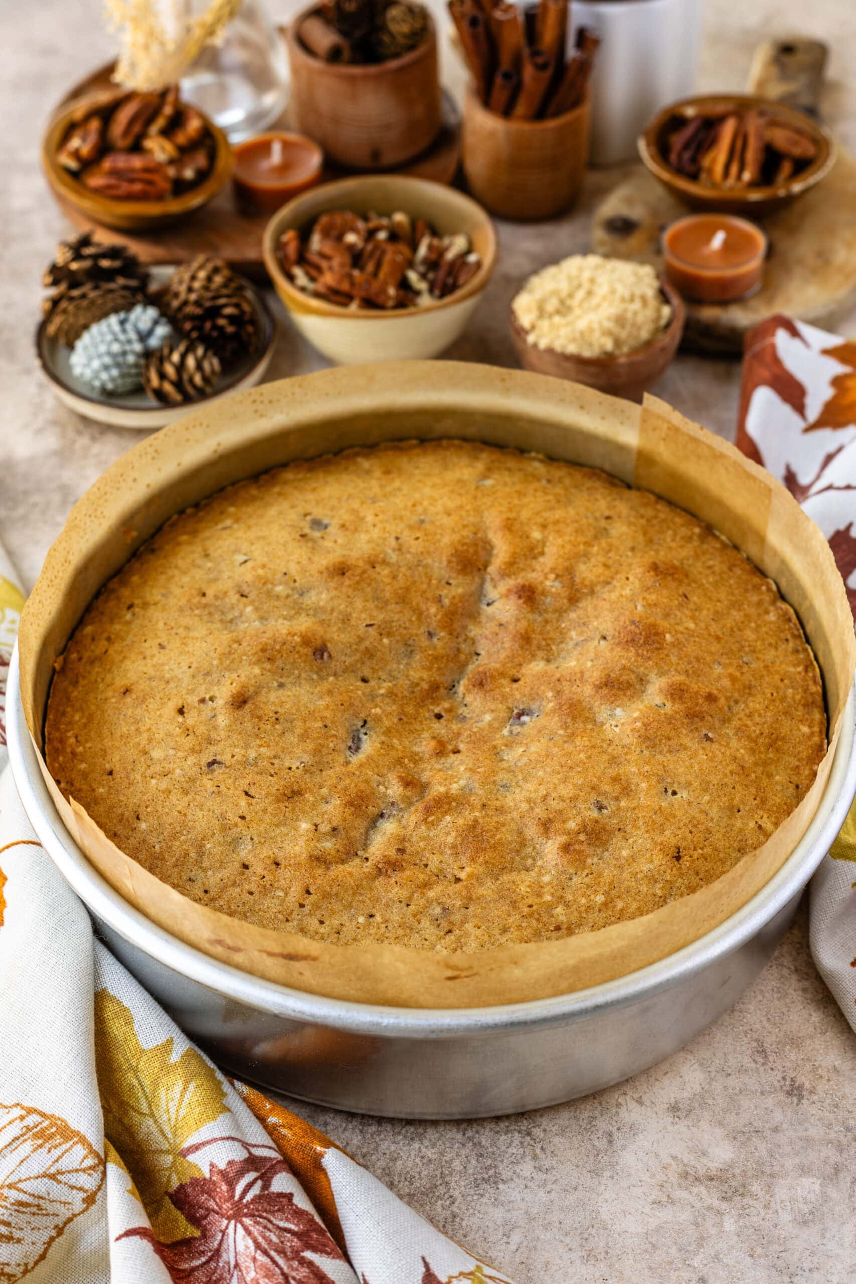 The image shows the freshly baked pecan pie cake, still in its round cake pan lined with parchment paper. The top of the cake is golden brown and evenly baked, with a slight rise, indicating a perfectly moist texture inside. In the background, there are small bowls filled with pecans, brown sugar, and cinnamon sticks, along with candles and autumn-themed decorations that create a cozy, seasonal ambiance. The cake is ready to be removed from the pan and flipped to reveal the caramelized pecan topping underneath.