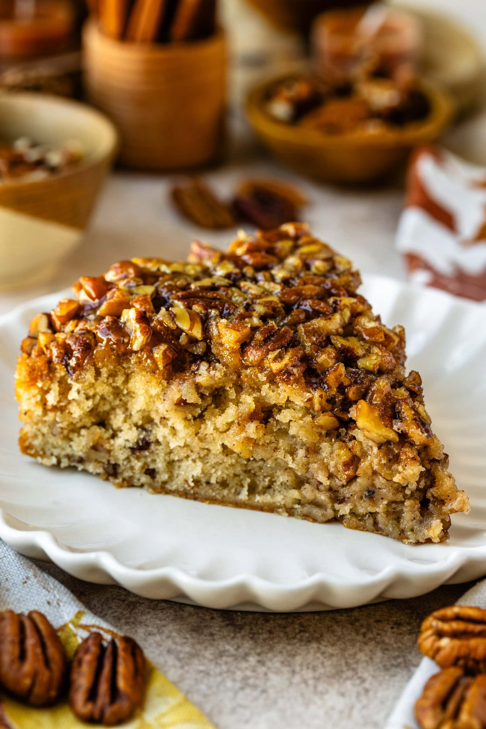 The image shows a close-up slice of pecan pie cake on a white scalloped plate. The cake is golden and moist, topped generously with chopped pecans glazed with caramelized syrup, giving it a rich, glossy finish. In the background, small bowls filled with pecans and other autumnal decor can be seen, enhancing the warm, cozy feeling of the dessert setting. The texture of the cake looks soft and fluffy, contrasted with the crunchy, sweet pecan topping.