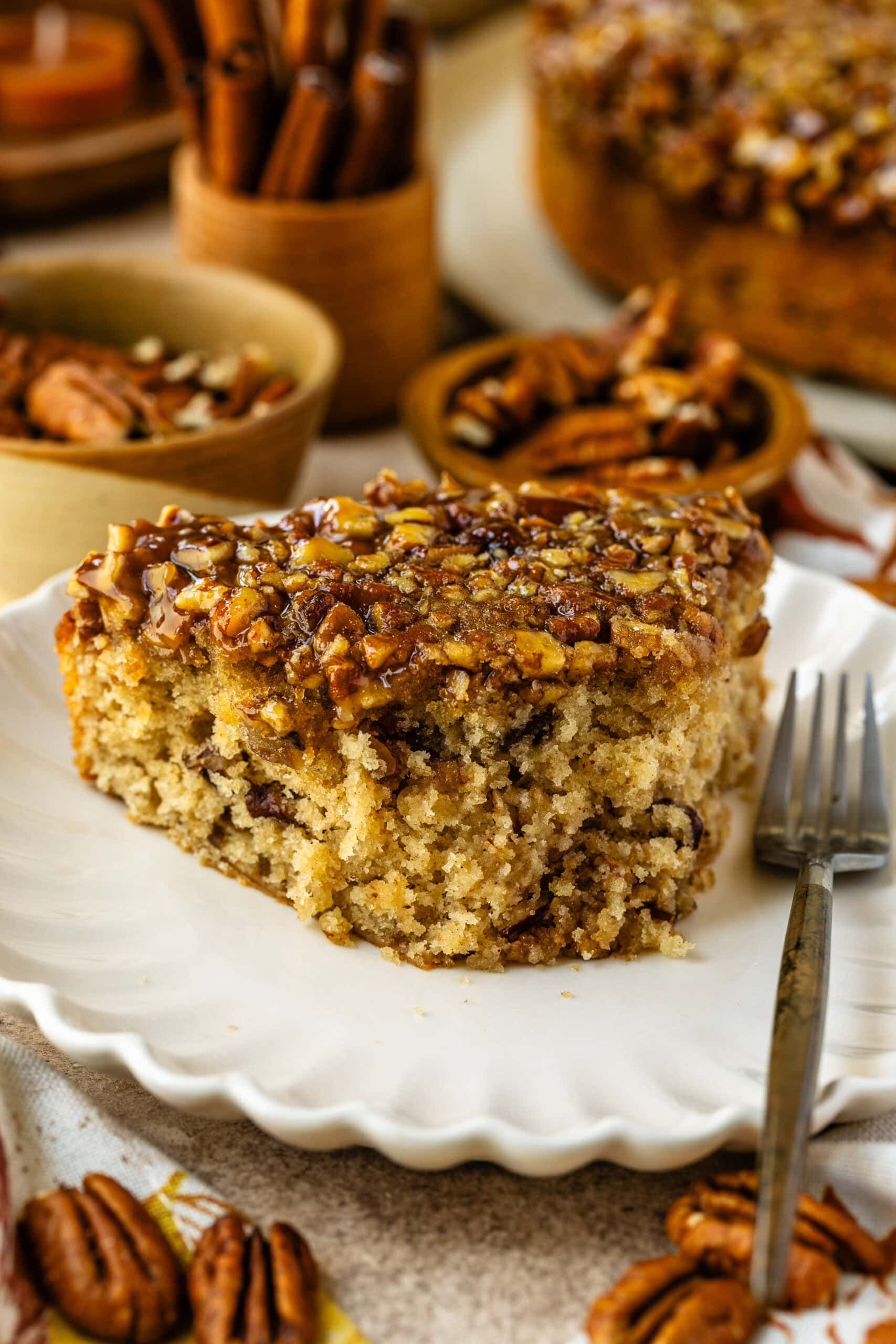 The image shows a close-up slice of pecan pie cake on a white scalloped plate. The cake is golden and moist, topped generously with chopped pecans glazed with caramelized syrup, giving it a rich, glossy finish. In the background, small bowls filled with pecans and other autumnal decor can be seen, enhancing the warm, cozy feeling of the dessert setting. The texture of the cake looks soft and fluffy, contrasted with the crunchy, sweet pecan topping.