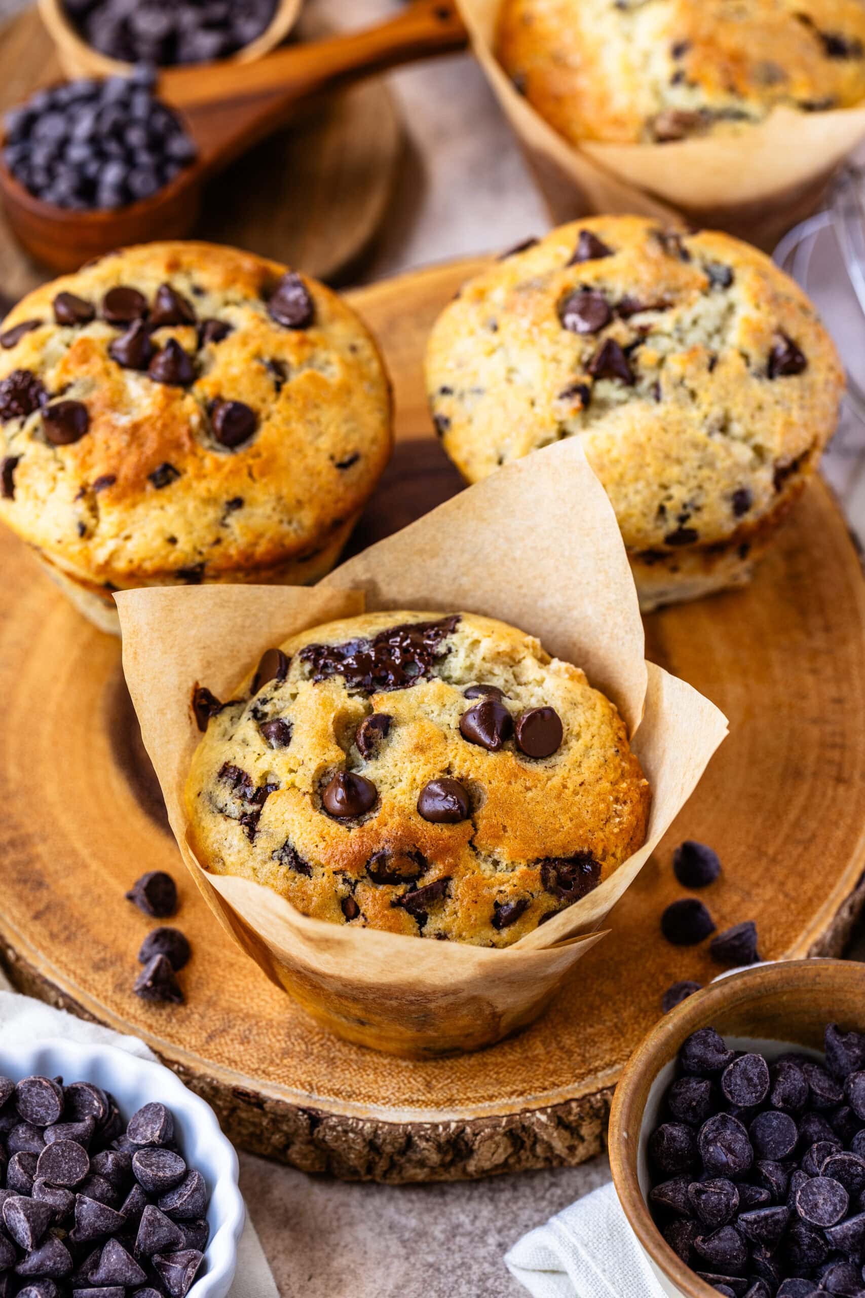 This image displays a beautifully arranged batch of freshly baked chocolate chip muffins on a rustic wooden platter. The muffins are golden brown, with plenty of chocolate chips scattered on top, some melted into the batter, adding to their decadent appearance. Each muffin is wrapped in tulip-style parchment paper liners, which give them a charming, bakery-style presentation. Surrounding the muffins are small bowls filled with extra chocolate chips, enhancing the indulgent appeal of these delicious treats. The scene is warm, inviting, and perfect for showcasing these homemade muffins.