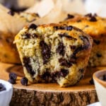 The close-up image features a chocolate chip muffin cut in half, revealing its moist and fluffy interior packed with melted chocolate chips. The rich chocolate is swirled throughout the soft, tender crumb of the muffin, making it look irresistibly delicious. The golden-brown outer crust contrasts beautifully with the warm, chocolatey center. In the background, more muffins in parchment liners are visible, creating an inviting scene that highlights the decadent texture and flavor of these homemade treats.