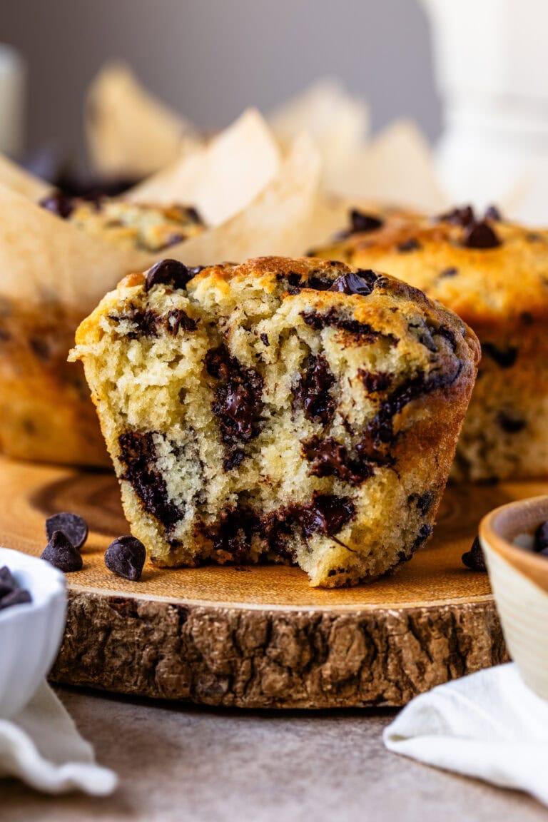 The close-up image features a chocolate chip muffin cut in half, revealing its moist and fluffy interior packed with melted chocolate chips. The rich chocolate is swirled throughout the soft, tender crumb of the muffin, making it look irresistibly delicious. The golden-brown outer crust contrasts beautifully with the warm, chocolatey center. In the background, more muffins in parchment liners are visible, creating an inviting scene that highlights the decadent texture and flavor of these homemade treats.