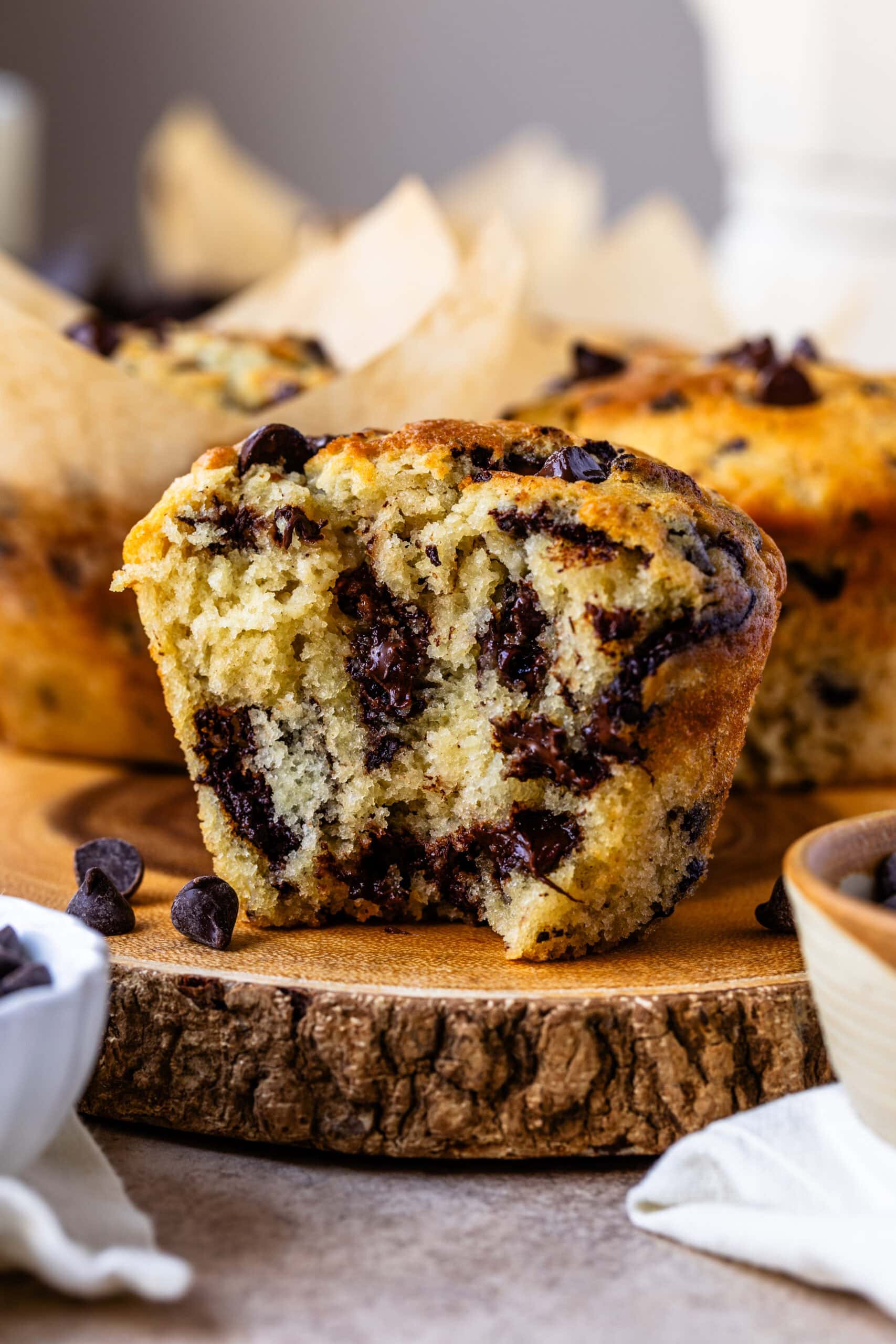 The close-up image features a chocolate chip muffin cut in half, revealing its moist and fluffy interior packed with melted chocolate chips. The rich chocolate is swirled throughout the soft, tender crumb of the muffin, making it look irresistibly delicious. The golden-brown outer crust contrasts beautifully with the warm, chocolatey center. In the background, more muffins in parchment liners are visible, creating an inviting scene that highlights the decadent texture and flavor of these homemade treats.