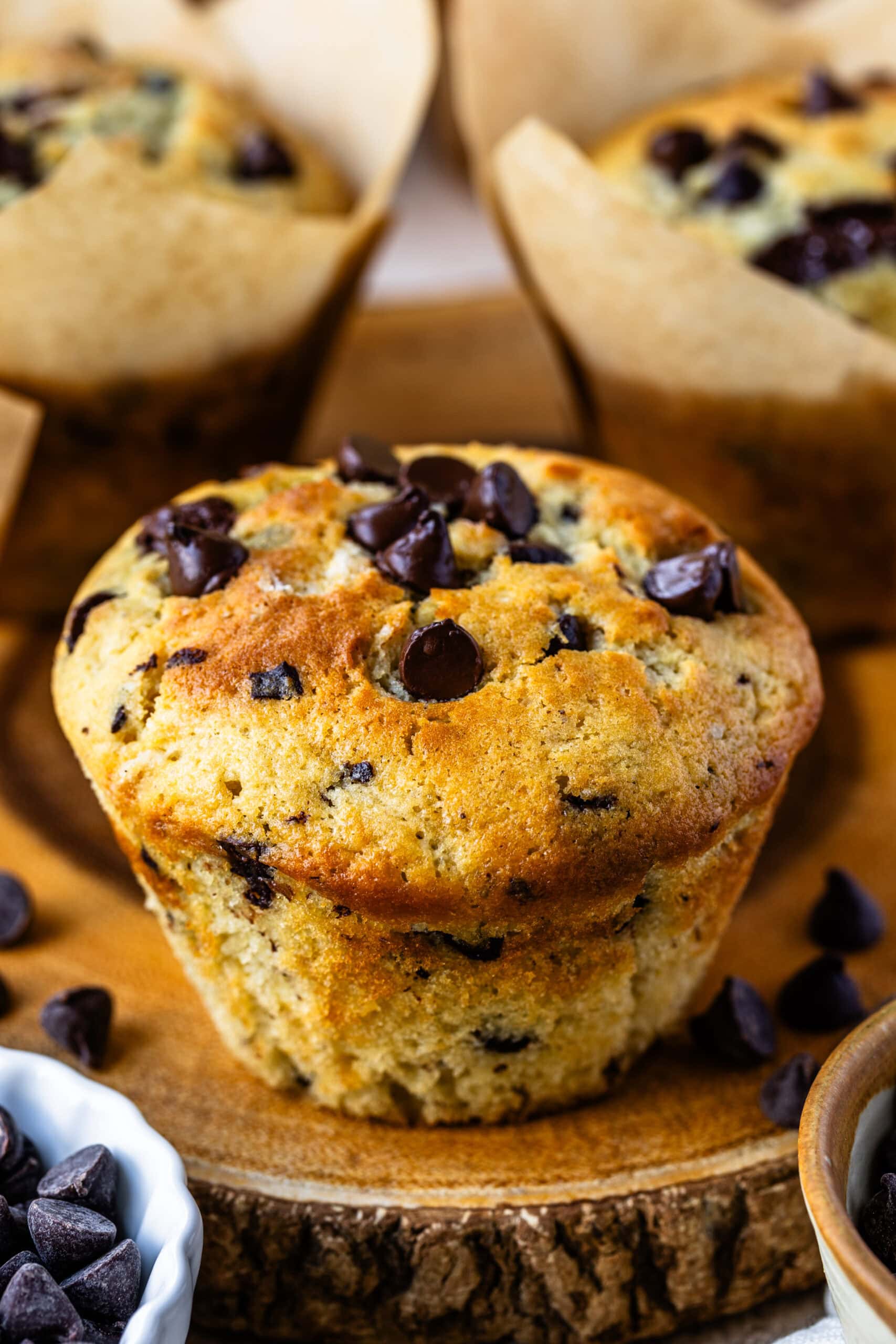 The close-up image features a single, beautifully baked chocolate chip muffin placed on a rustic wooden surface. The muffin has a golden-brown top with a generous sprinkle of chocolate chips, which are slightly melted, giving it a rich and indulgent appearance. The texture of the muffin looks soft and fluffy, with chocolate chunks visible throughout. The background showcases additional muffins in parchment liners, creating a warm and inviting scene perfect for highlighting the deliciousness of this homemade treat.