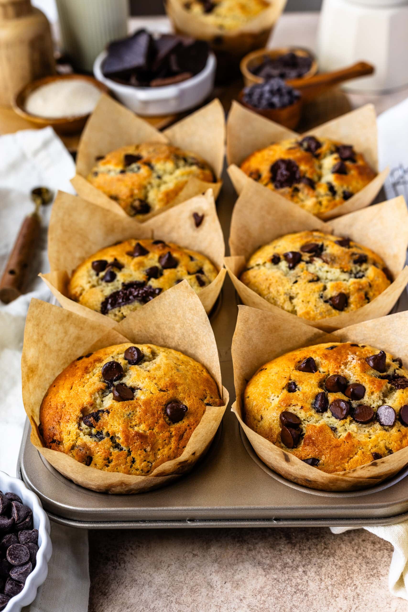 The image shows freshly baked chocolate chip muffins, golden brown and perfectly risen, still in their tulip-style parchment paper liners in a muffin tin. The tops are studded with melty chocolate chips, adding a delicious contrast to the fluffy texture of the muffins. The background includes ingredients like chocolate chunks and sugar in small bowls, creating a warm and inviting atmosphere that highlights the homemade charm of these delightful treats.