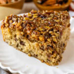 The image shows a close-up slice of pecan pie cake on a white scalloped plate. The cake is golden and moist, topped generously with chopped pecans glazed with caramelized syrup, giving it a rich, glossy finish. In the background, small bowls filled with pecans and other autumnal decor can be seen, enhancing the warm, cozy feeling of the dessert setting. The texture of the cake looks soft and fluffy, contrasted with the crunchy, sweet pecan topping.