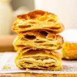 This close-up image showcases beautifully baked puff pastry, stacked to reveal the intricate, golden layers inside. The light, flaky texture is visible, with crisp edges and airy pockets throughout, perfectly demonstrating the lamination process that creates the signature puff. The pastry sits on a wooden board with a rustic, textured surface, highlighting the delicate craftsmanship of the baking process. The warm tones and natural lighting create a tempting and inviting presentation.