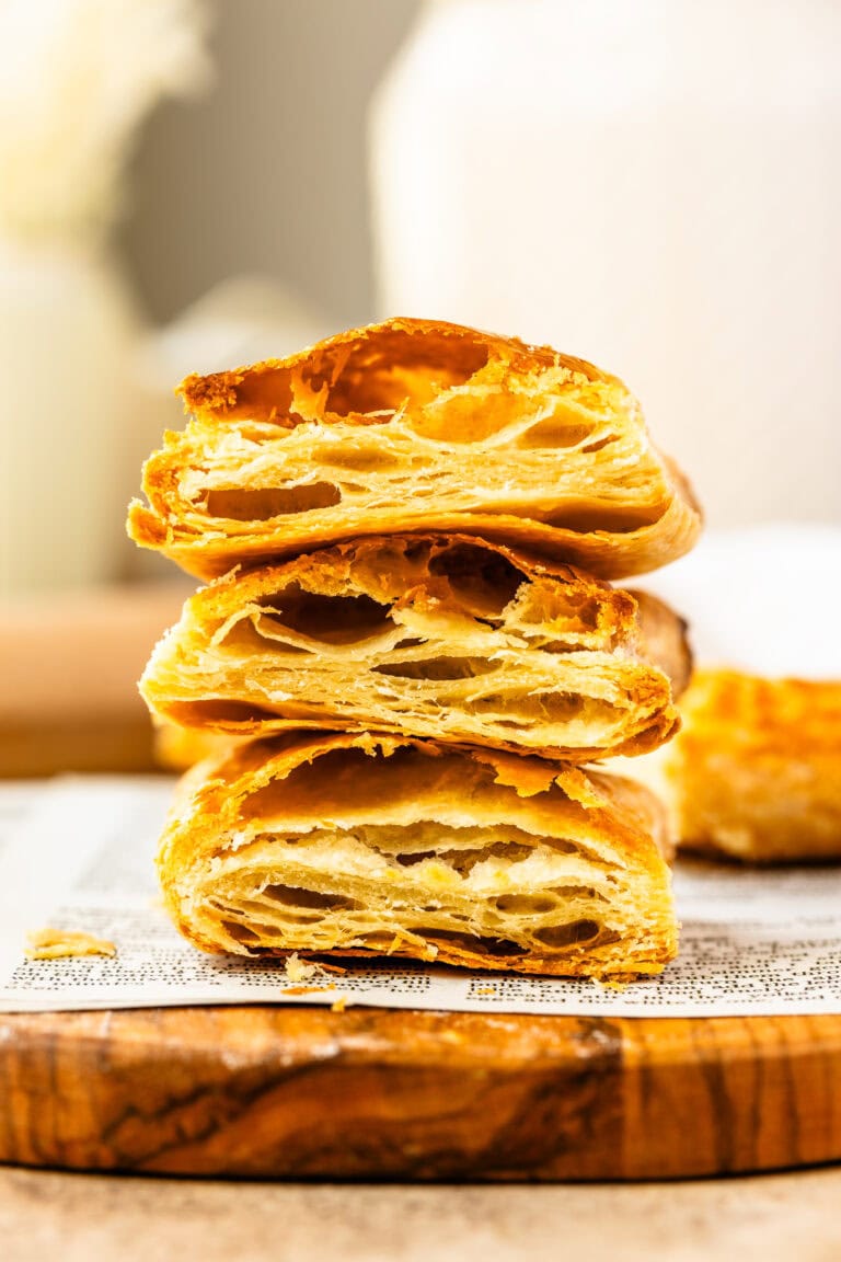 This close-up image showcases beautifully baked puff pastry, stacked to reveal the intricate, golden layers inside. The light, flaky texture is visible, with crisp edges and airy pockets throughout, perfectly demonstrating the lamination process that creates the signature puff. The pastry sits on a wooden board with a rustic, textured surface, highlighting the delicate craftsmanship of the baking process. The warm tones and natural lighting create a tempting and inviting presentation.