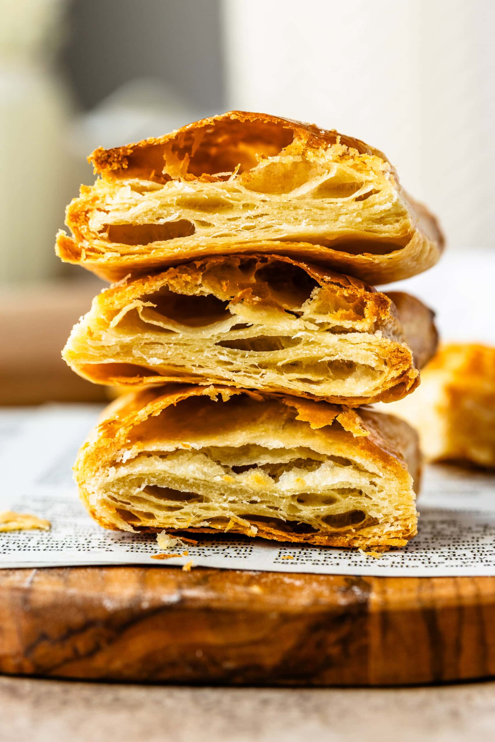 This close-up image showcases beautifully baked puff pastry, stacked to reveal the intricate, golden layers inside. The light, flaky texture is visible, with crisp edges and airy pockets throughout, perfectly demonstrating the lamination process that creates the signature puff. The pastry sits on a wooden board with a rustic, textured surface, highlighting the delicate craftsmanship of the baking process. The warm tones and natural lighting create a tempting and inviting presentation.
