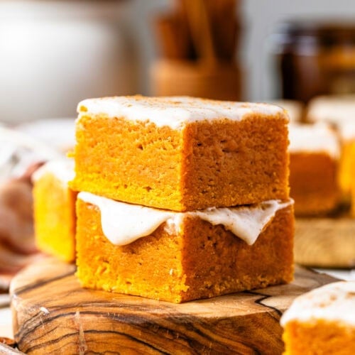 Two stacked pumpkin blondie bars, topped with a creamy frosting, placed on a wooden board. The blondies have a rich, golden-orange color and a moist texture, with the frosting glistening under soft lighting. The warm, blurred background adds to the inviting, fall-themed atmosphere.