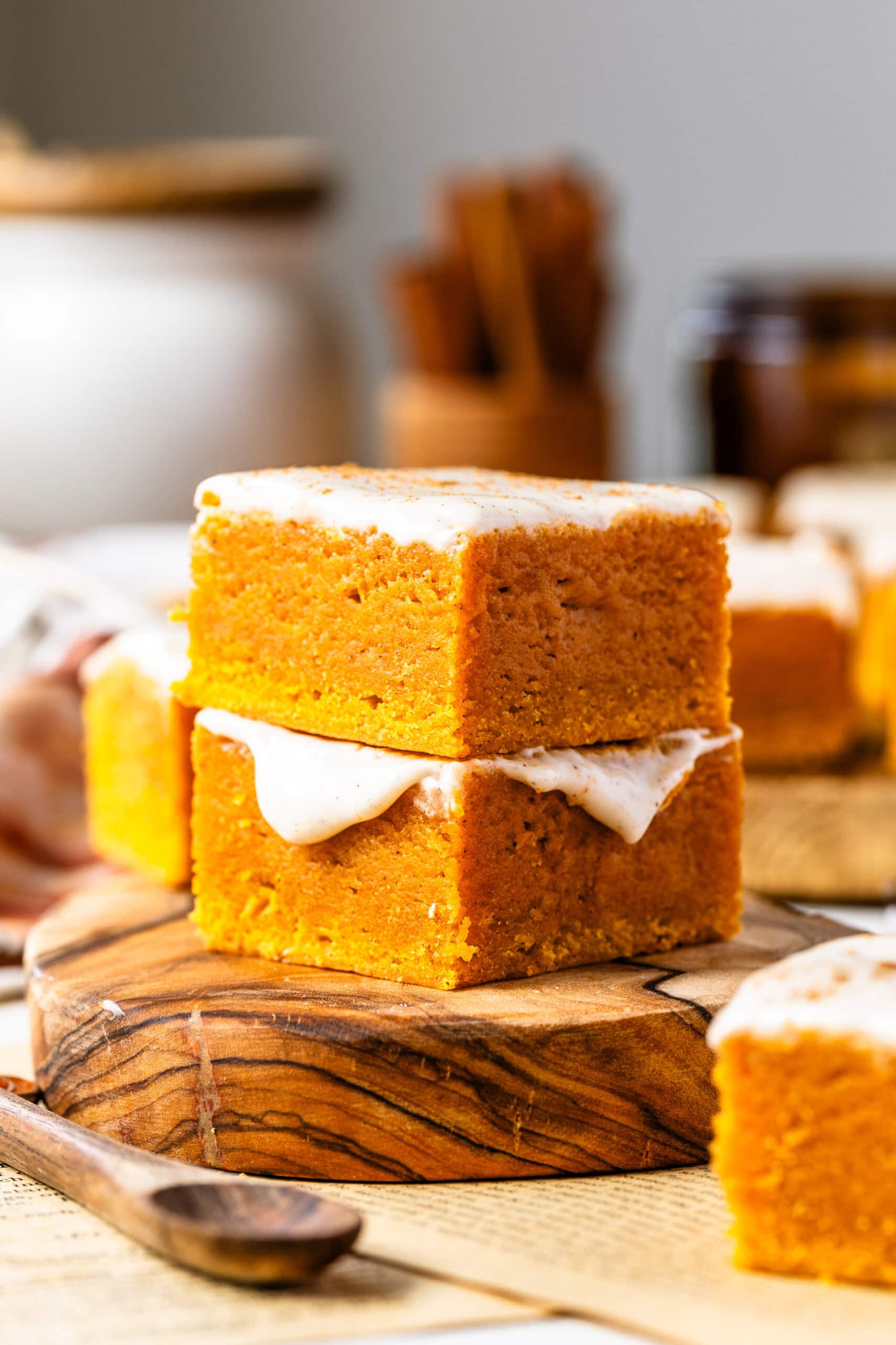 Two stacked pumpkin blondie bars, topped with a creamy frosting, placed on a wooden board. The blondies have a rich, golden-orange color and a moist texture, with the frosting glistening under soft lighting. The warm, blurred background adds to the inviting, fall-themed atmosphere.