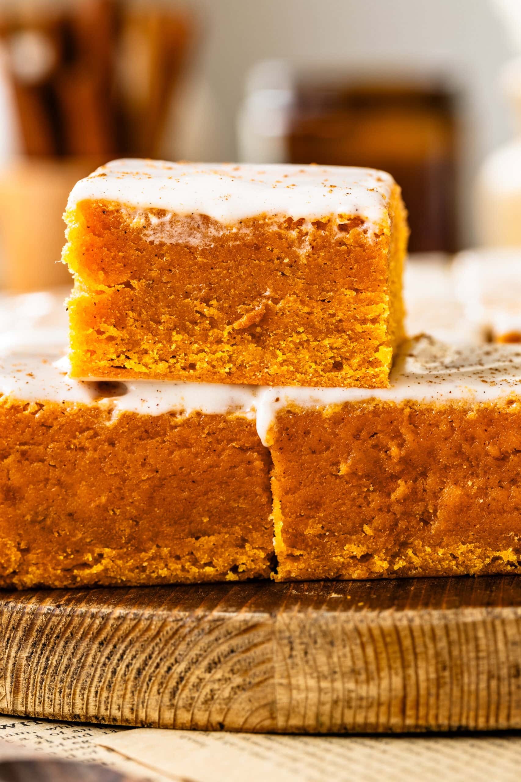 Image features a close-up of a single pumpkin blondie piece stacked on another, emphasizing the dense, moist crumb and creamy frosting on top. The wooden surface adds a rustic touch, and the blurred background enhances the cozy, seasonal feel.