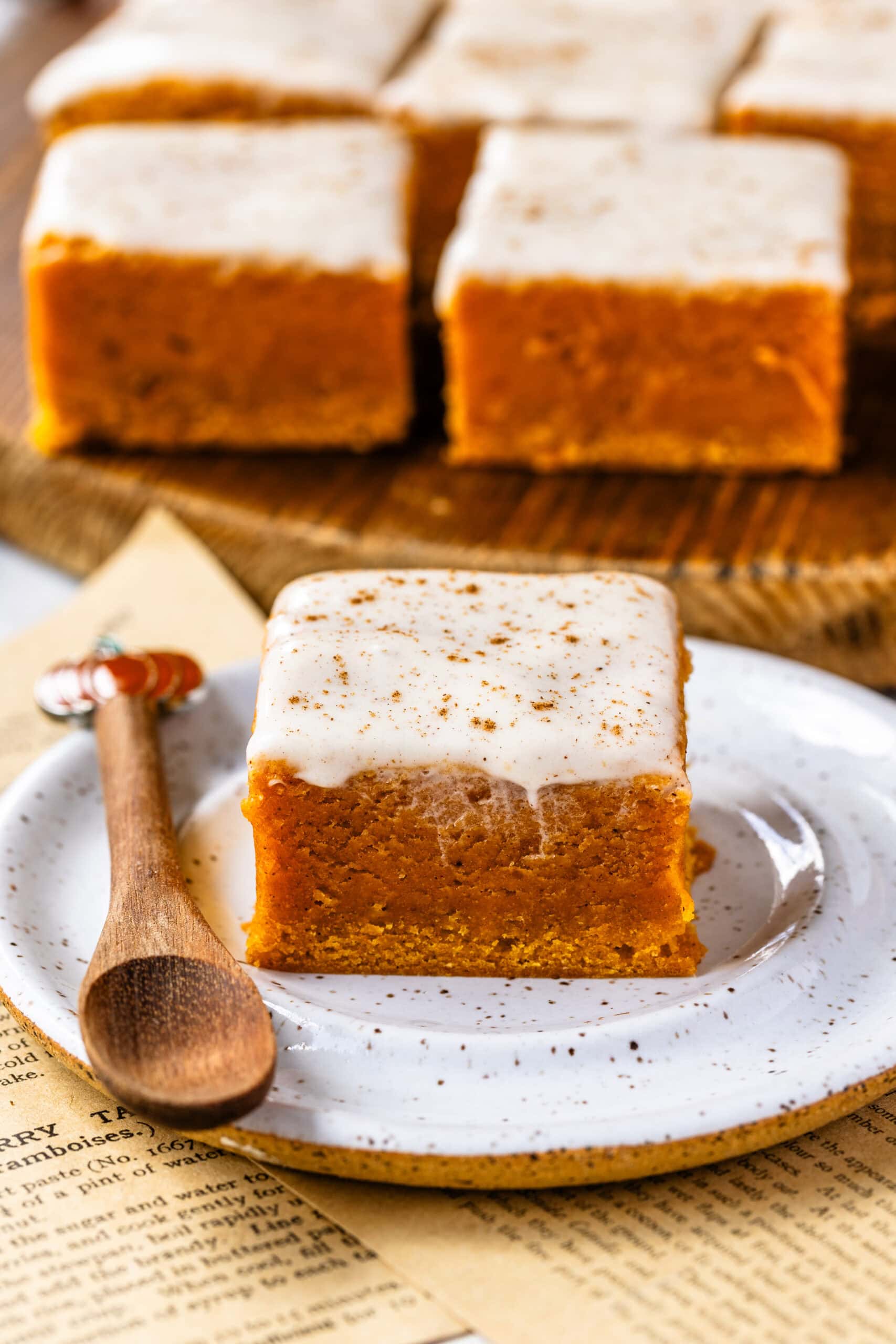 A close-up image of a single pumpkin blondie square topped with creamy glaze, sitting on a white speckled plate with a small wooden spoon beside it. The blondie's soft, dense texture and glossy frosting are beautifully highlighted, with a sprinkle of spices on top, while blurred blondies in the background add to the warm, inviting autumn feel.