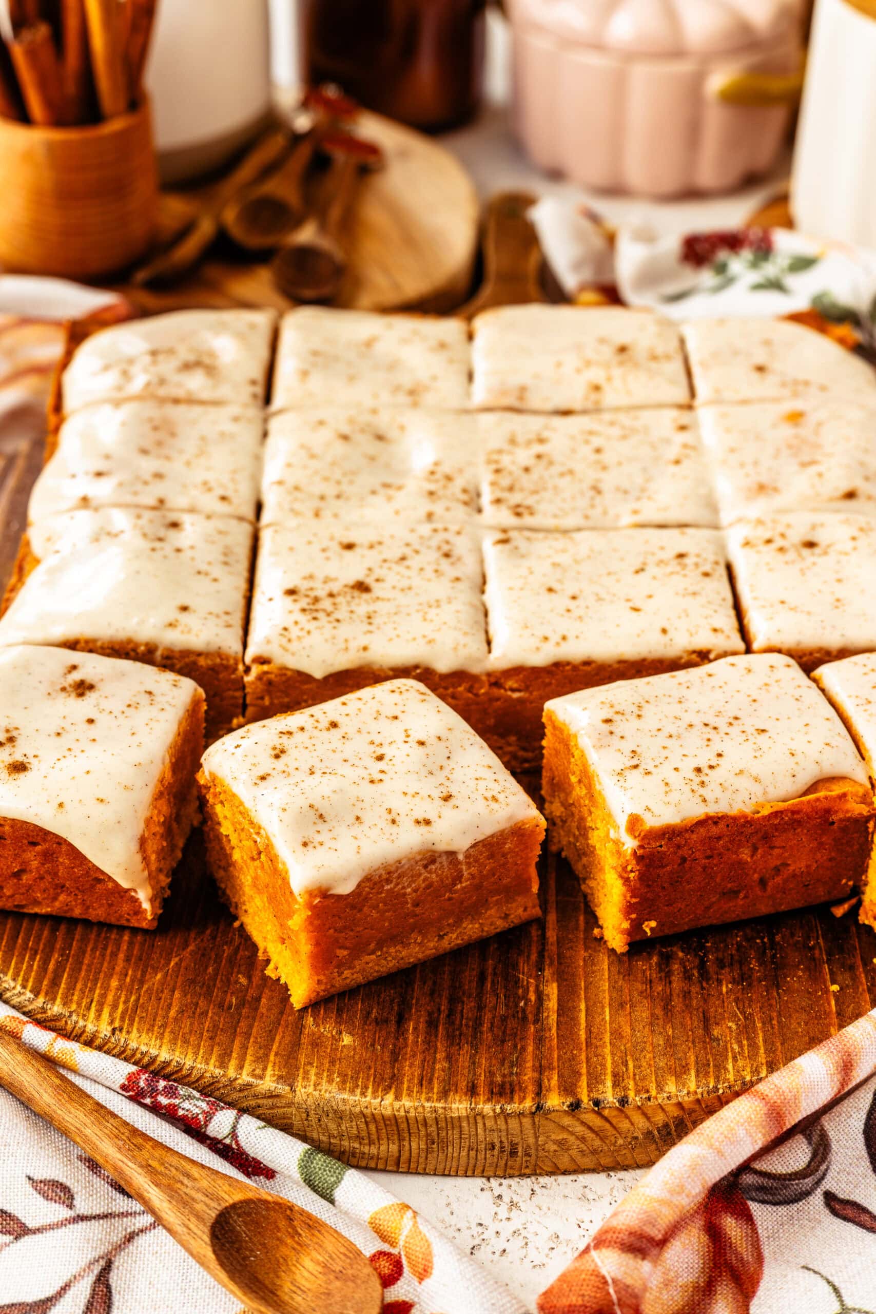 A batch of pumpkin blondies cut into squares, topped with a creamy, spiced glaze, and beautifully arranged on a wooden board. Two blondie squares are placed in the foreground, showcasing their moist texture and smooth glaze, while the blurred background highlights autumn-themed decor. The wooden surface and floral napkin enhance the warm, inviting fall atmosphere of this delectable treat.