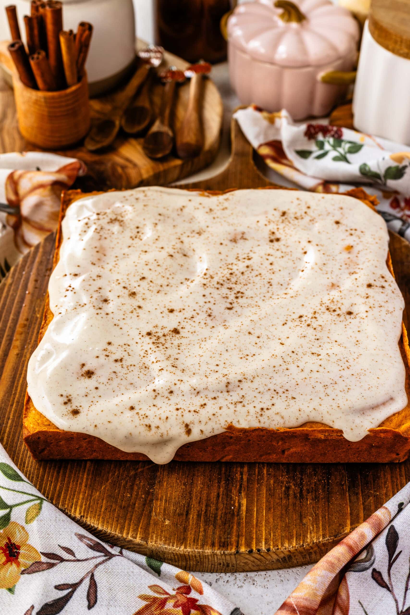 A freshly glazed pumpkin blondie block sits on a wooden board, topped with a thick layer of creamy glaze sprinkled with spices. The smooth frosting is generously spread over the blondie, with a few drips along the edges for a rustic touch. The warm fall-themed setting, complete with cinnamon sticks, wooden utensils, and a floral napkin, creates a cozy and inviting atmosphere for this autumn dessert.